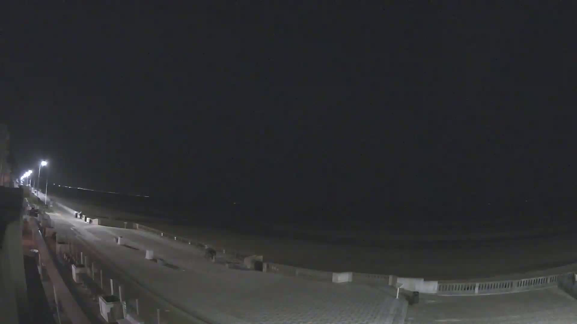 A nighttime view of a mostly empty beach boardwalk with a dark sky and calm ocean.