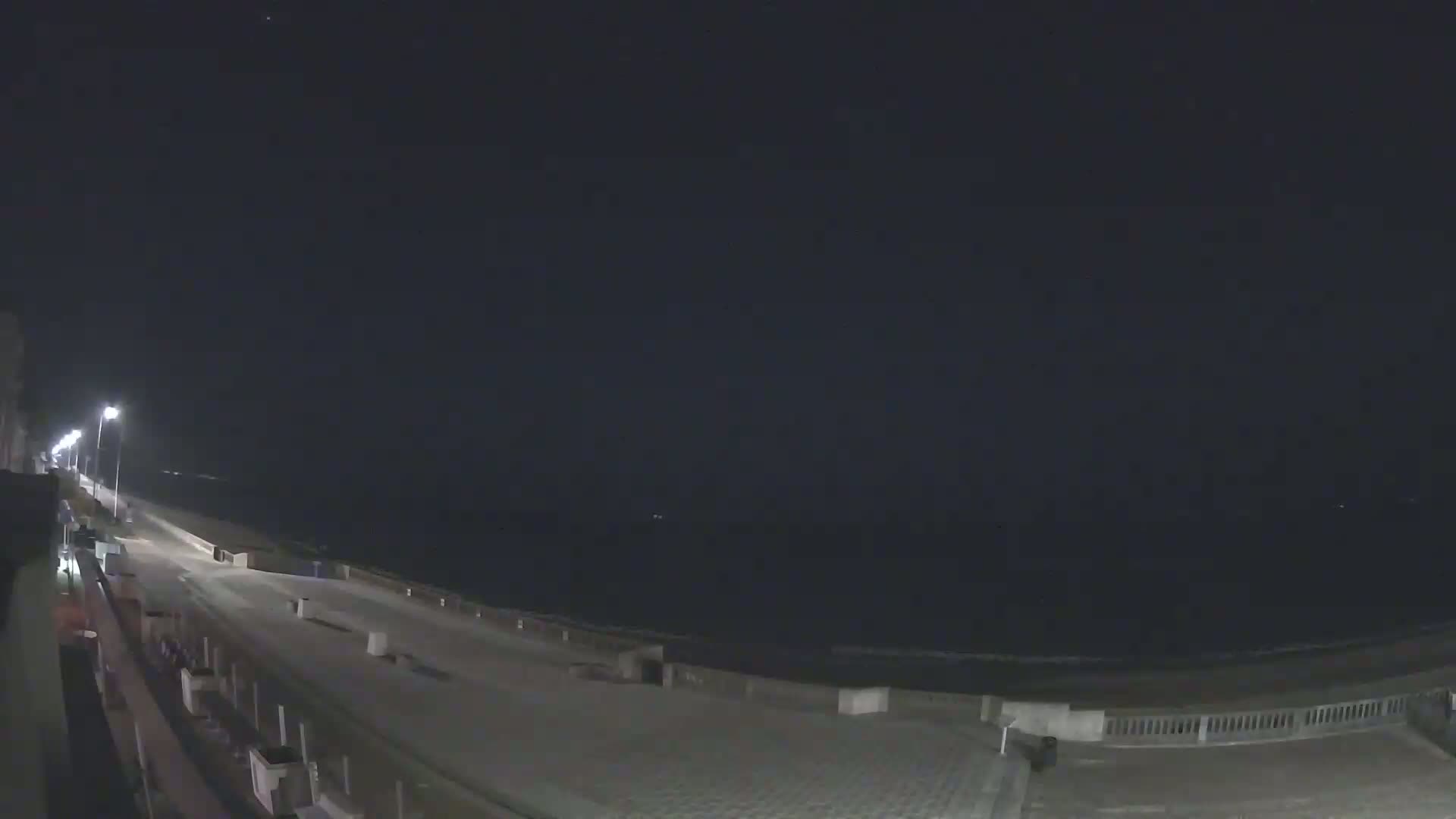 A nighttime view of a mostly empty boardwalk along a dark ocean under a clear sky.