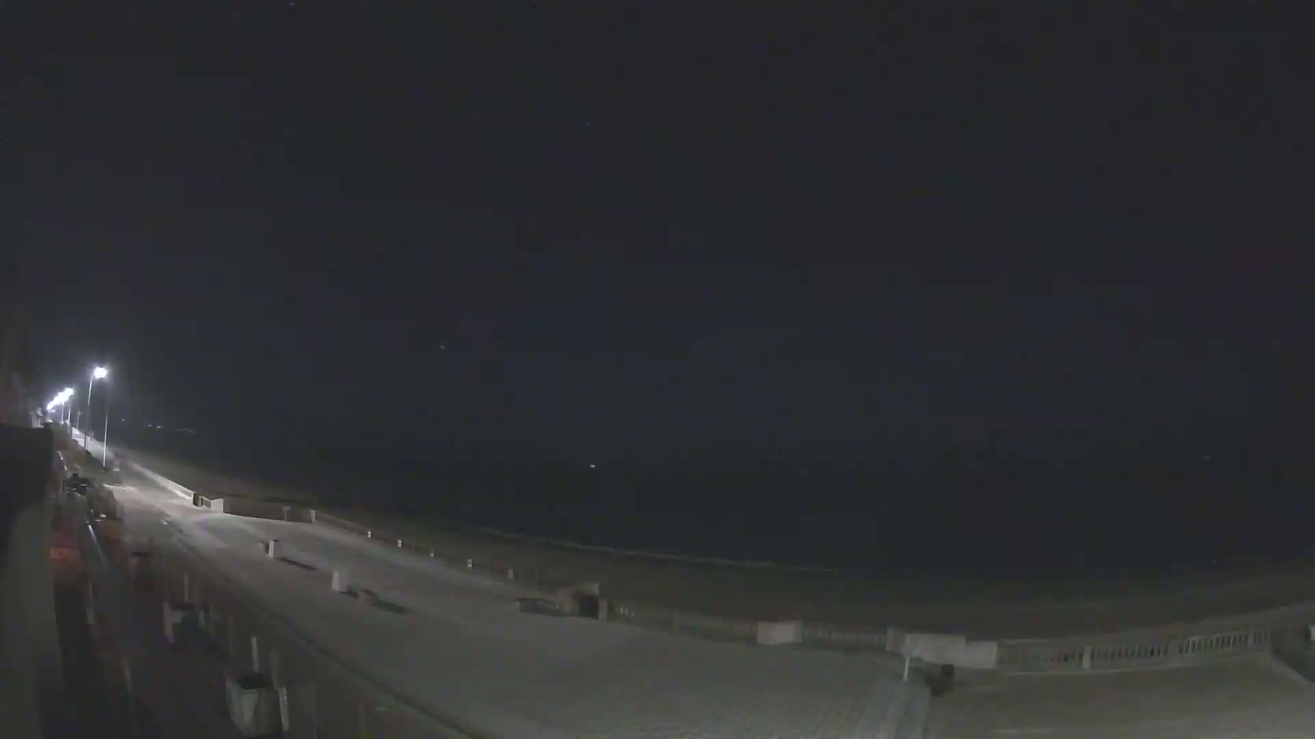A nighttime view of a mostly empty boardwalk and beach, illuminated by streetlights under a dark, clear sky.