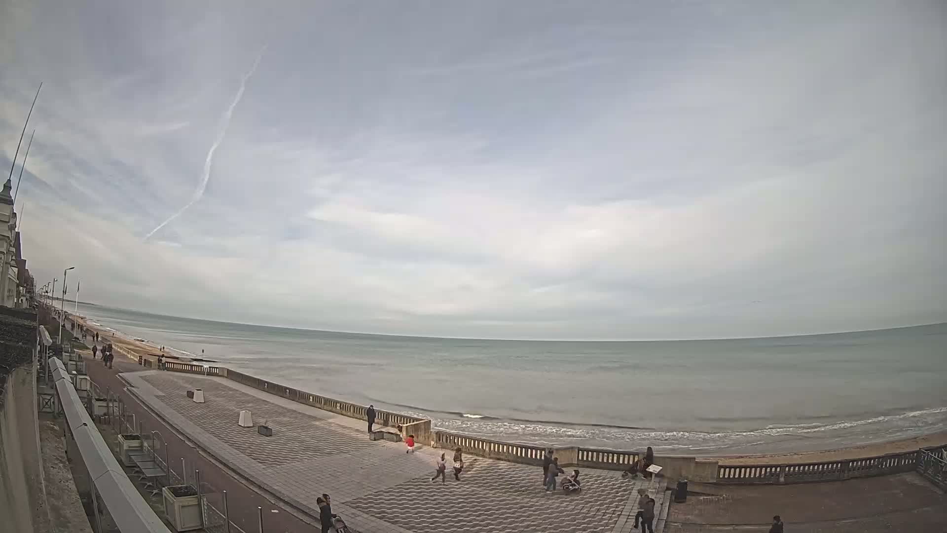 A wide, paved seaside promenade stretches alongside a calm, gray ocean under a mostly cloudy sky.