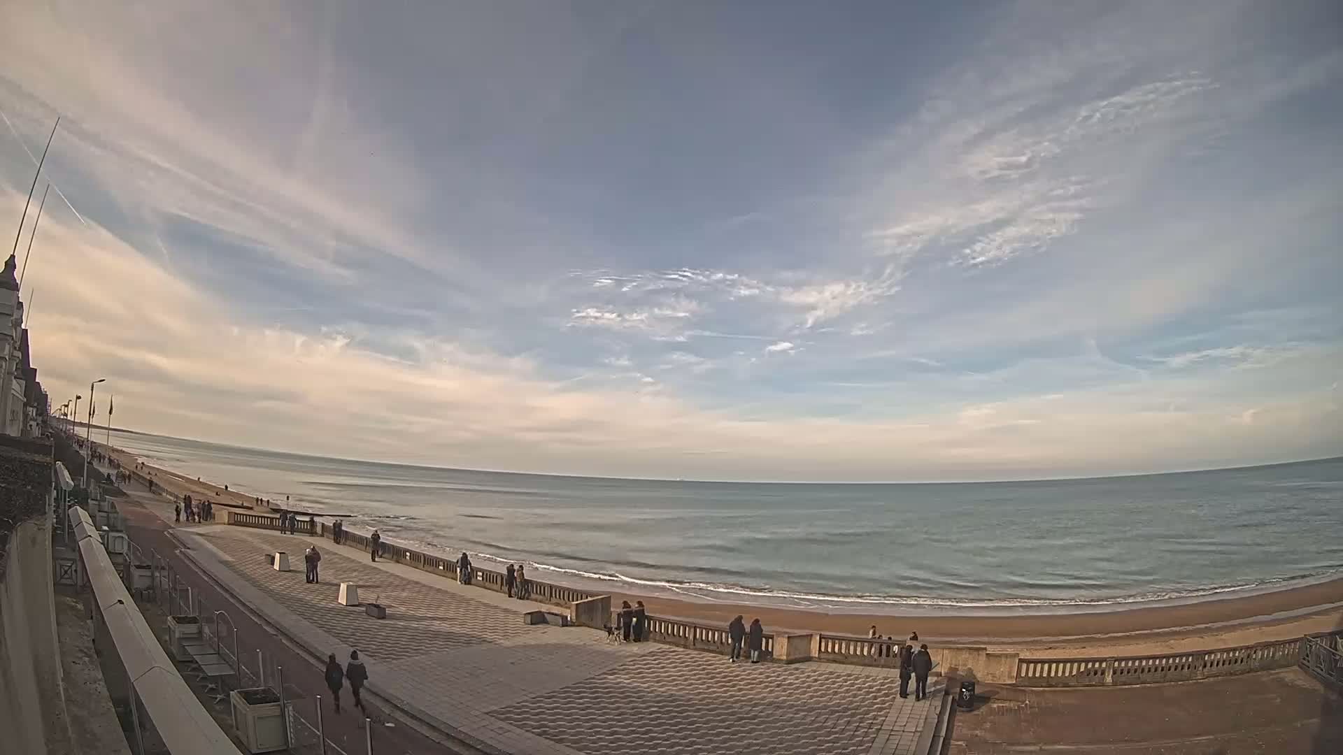 A mostly sunny day with scattered clouds shows a wide, sandy beach and calm ocean with people strolling along a paved walkway next to it.