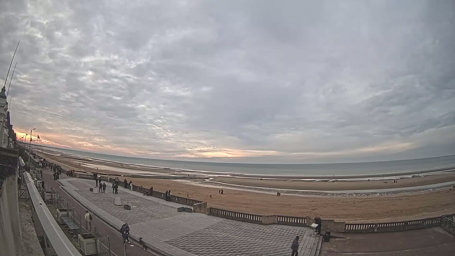 A wide shot shows a beach with people walking along the shore and a paved walkway at sunset under a mostly cloudy sky.