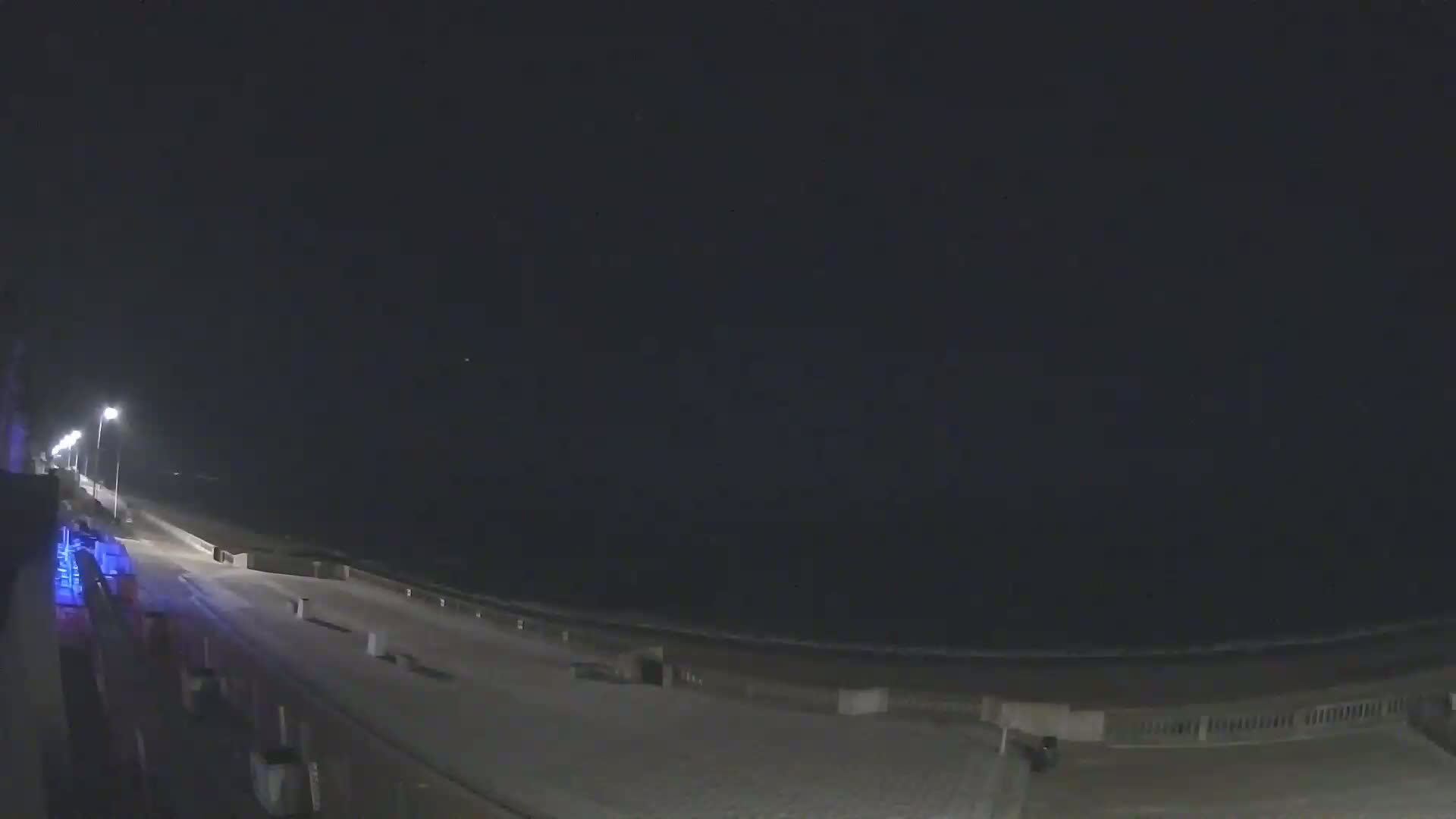 A nighttime view of a mostly empty boardwalk along a beach, lit by streetlights under a dark sky.