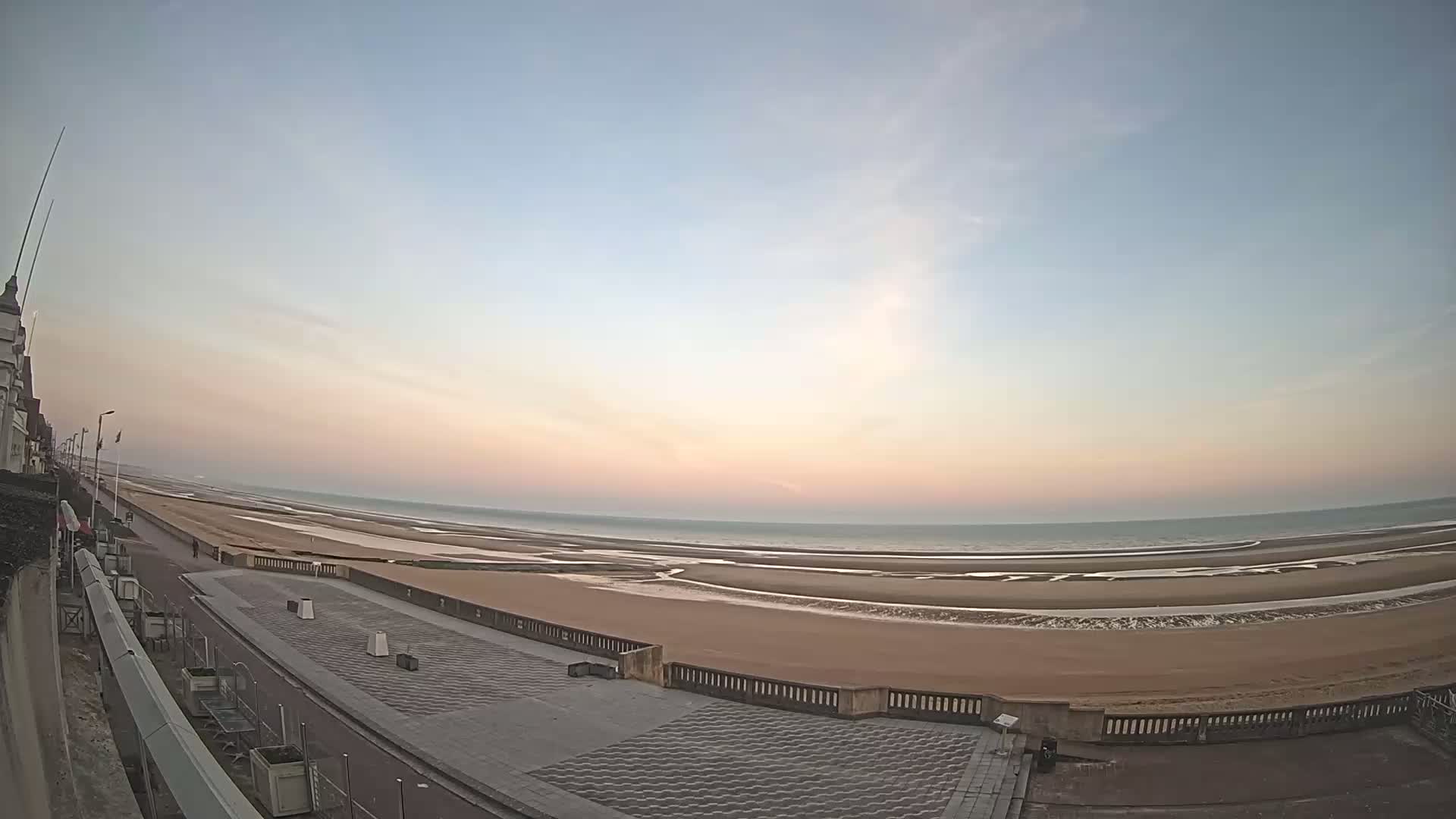 A wide shot of a sandy beach and promenade at sunrise, under a pale pink and blue sky.