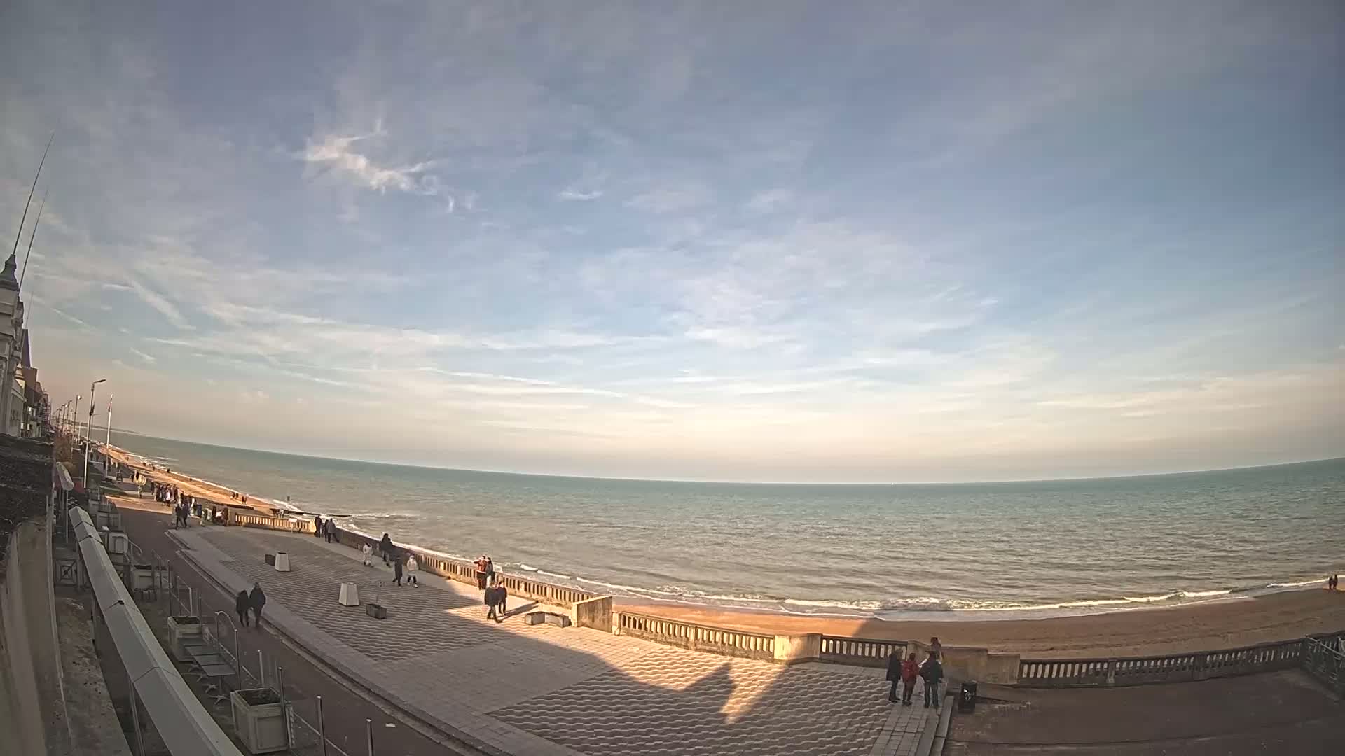 A mostly sunny day shows a wide expanse of calm ocean meeting a sandy beach, with a paved promenade and a few people walking along it.