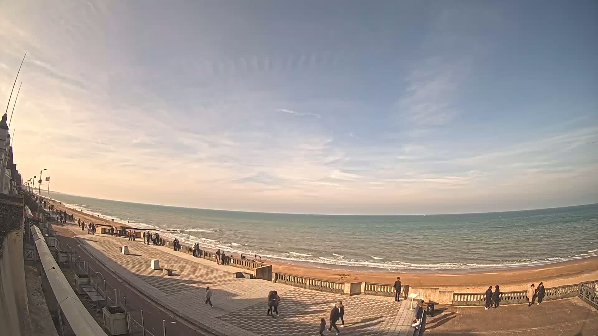 A wide shot shows a mostly sunny day at a beach with people walking along a paved walkway next to the sand and ocean.