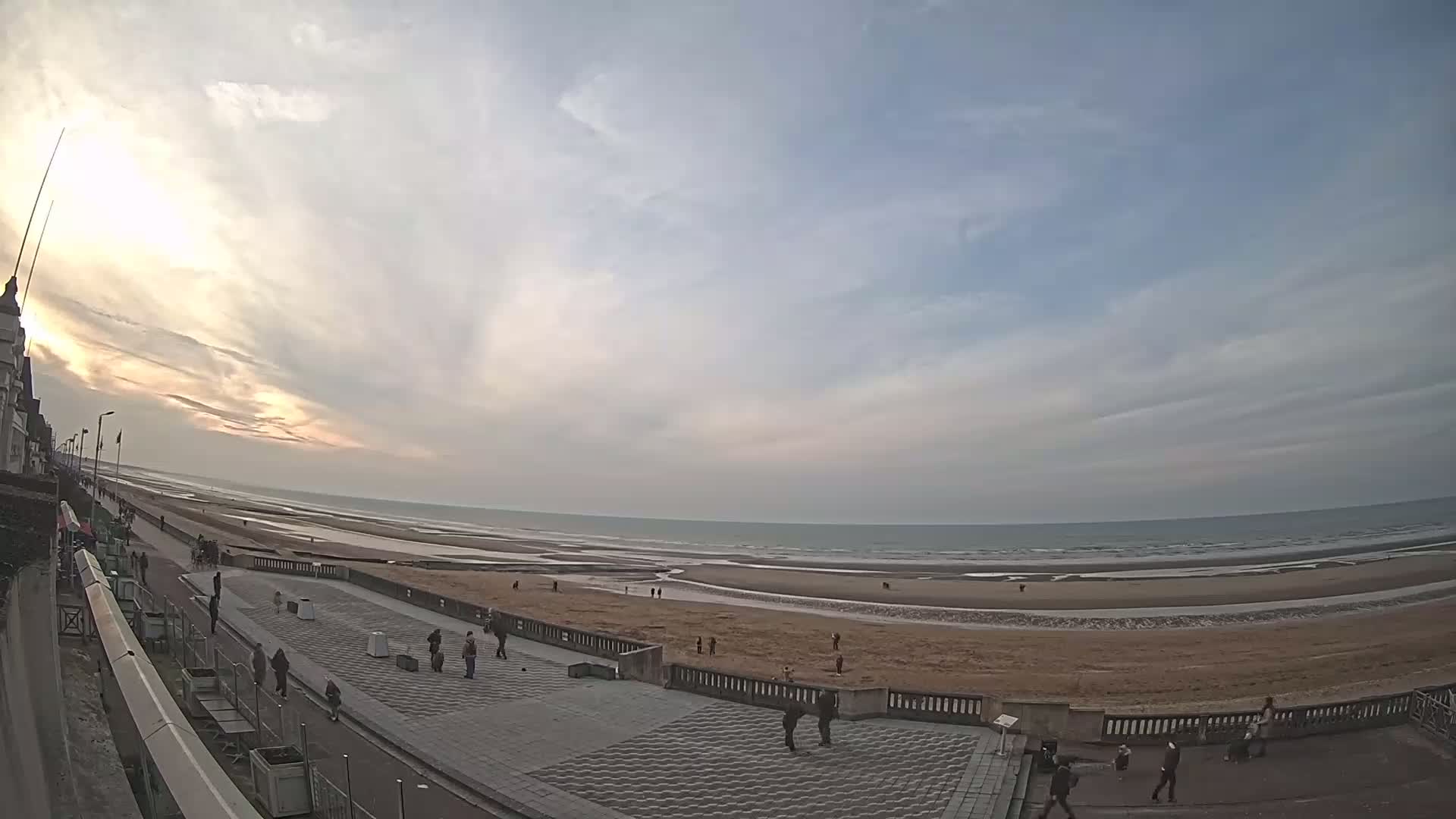 A wide shot shows a mostly cloudy sunset over a wide sandy beach and ocean, with a few people walking along the shoreline and paved promenade.