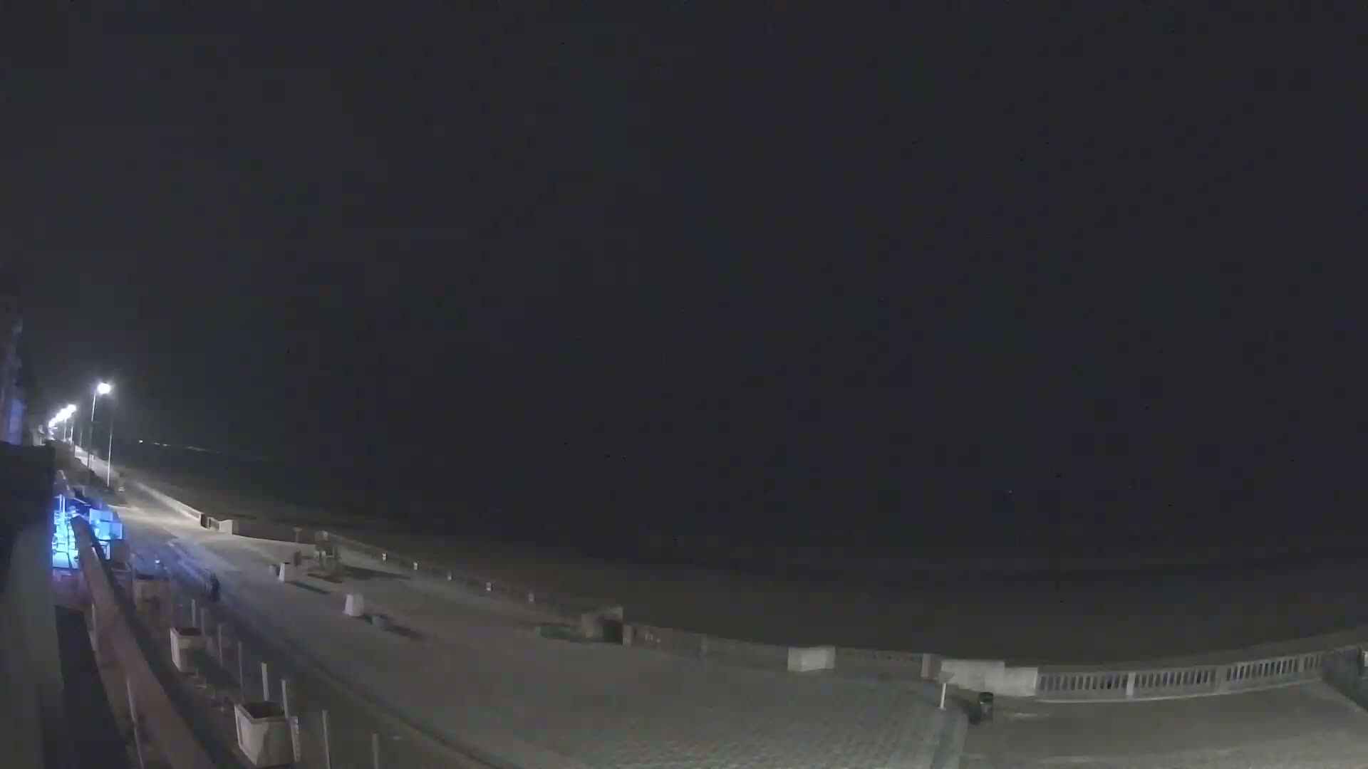 A nighttime view shows a snow-covered boardwalk along a dark ocean under a clear, black sky.