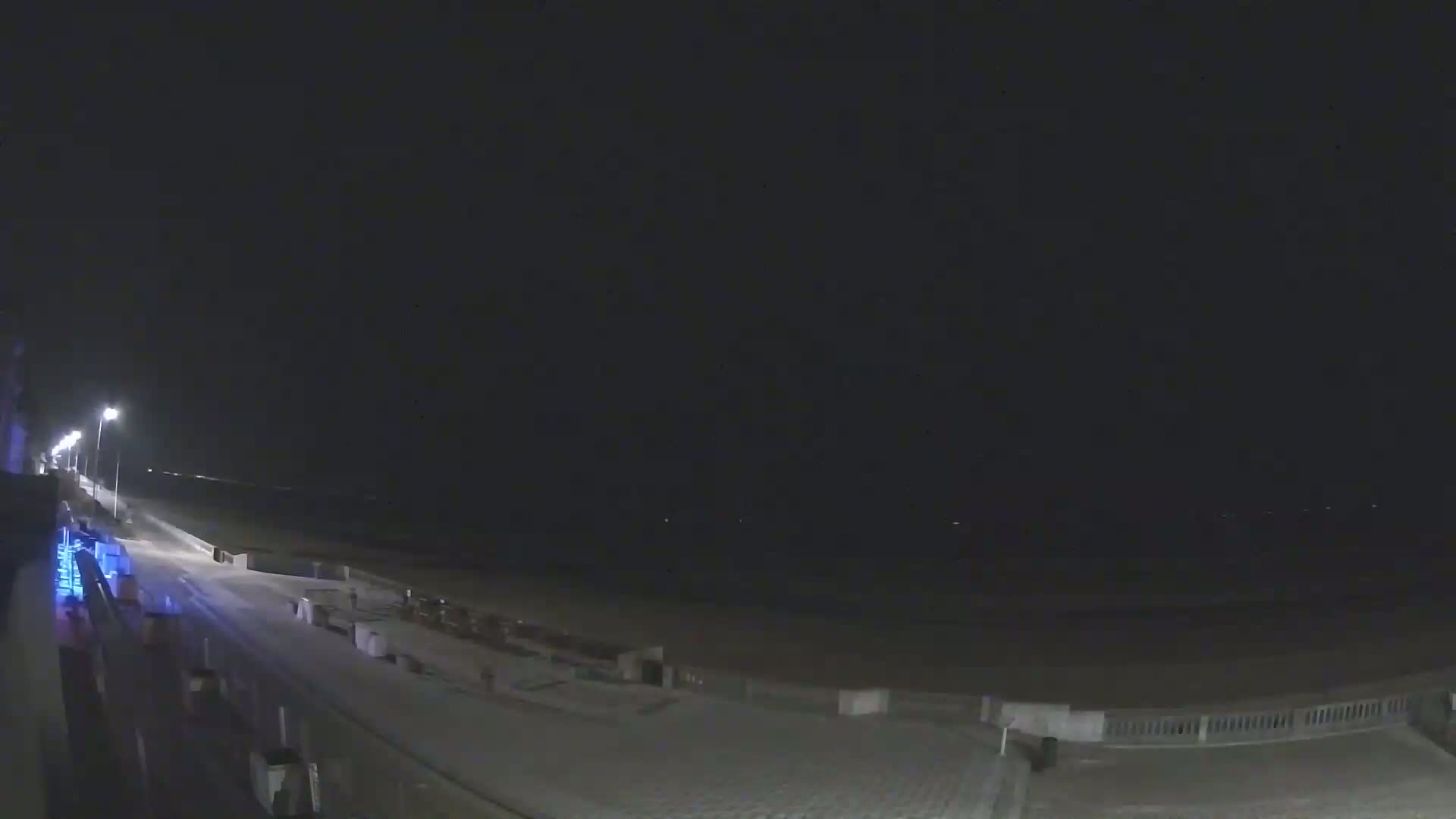 A nighttime view of a mostly empty beach boardwalk with streetlights illuminating a wet, dark beach and ocean under a cloudy sky.