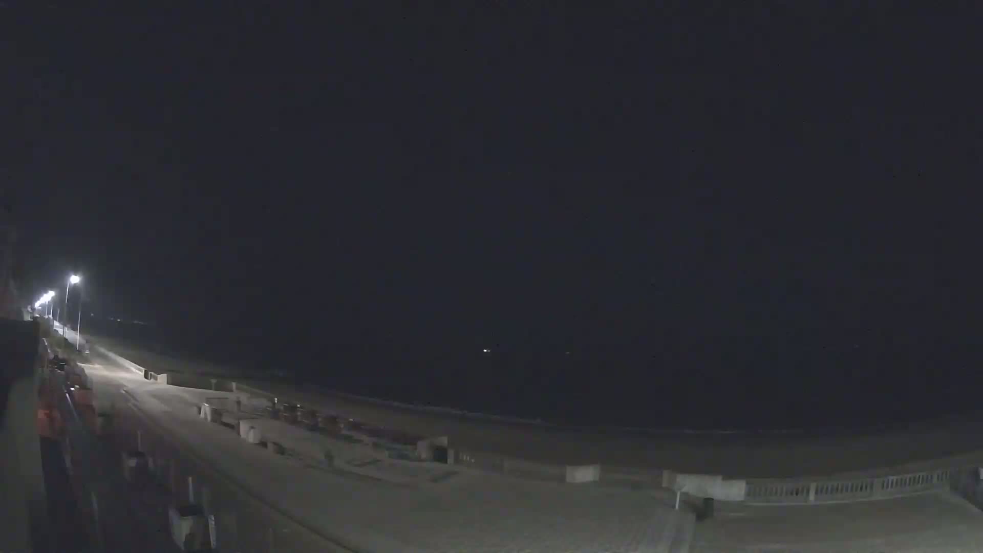 A nighttime view of a mostly empty beach and boardwalk with a few lights visible under a dark sky.