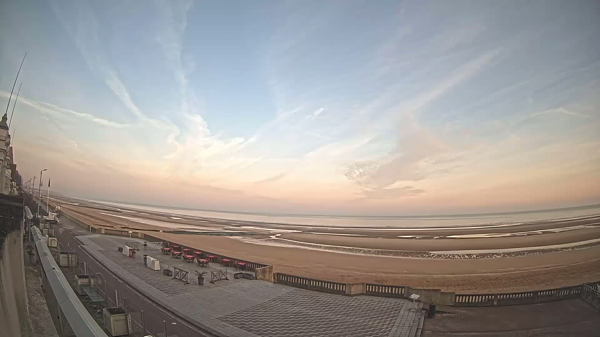A wide, panoramic view shows a sandy beach at low tide under a pale pink and blue sunrise sky with thin, wispy clouds.