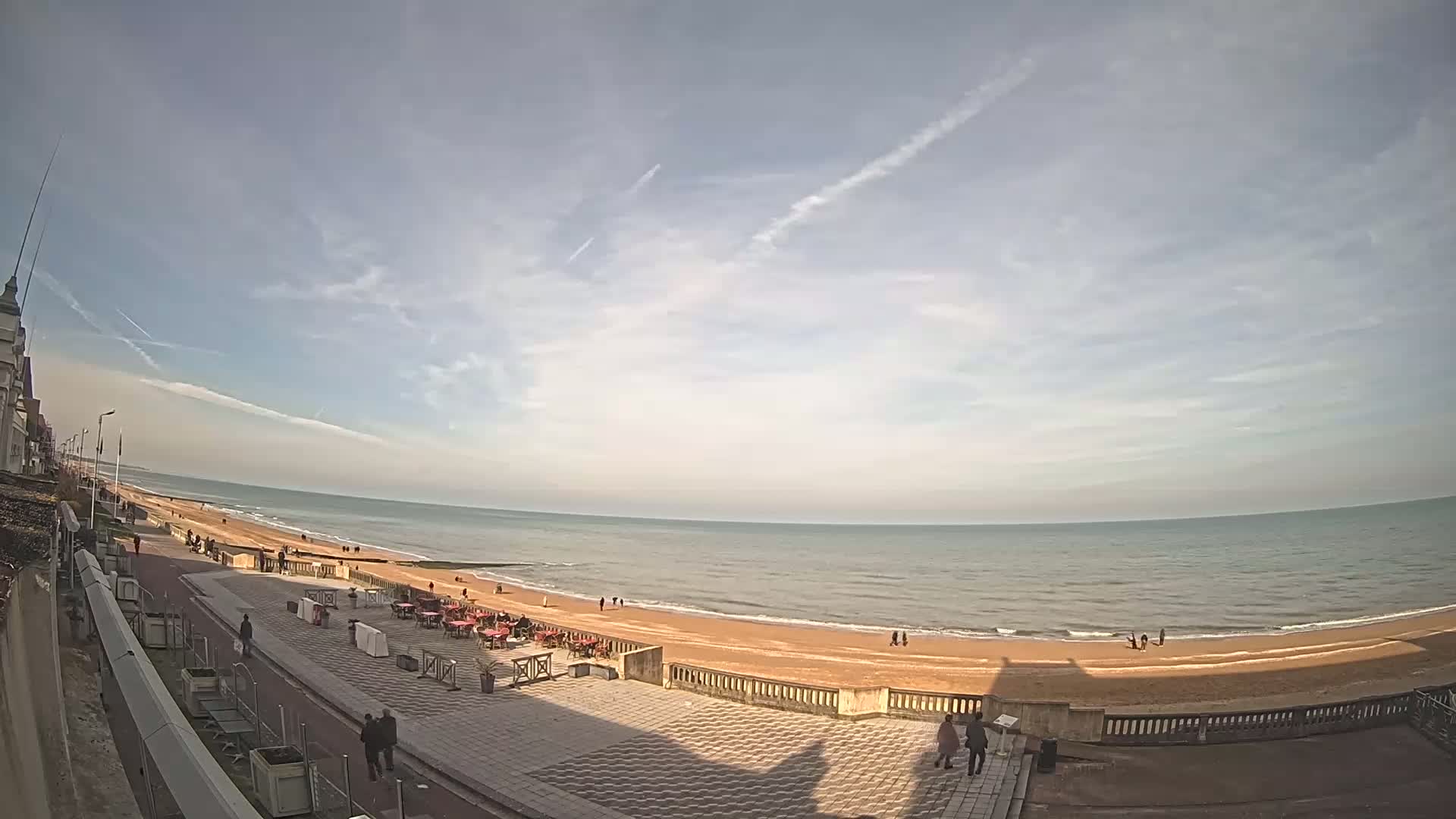 A partly cloudy day shows a wide expanse of sandy beach and calm ocean, with a promenade lined with outdoor seating and a few pedestrians.