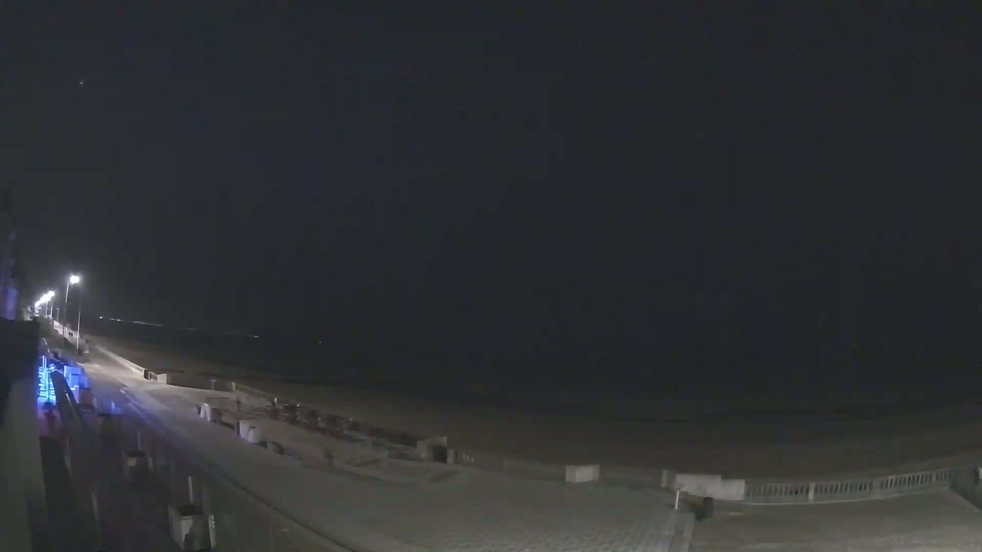 A nighttime view of a mostly empty beach boardwalk with streetlights illuminating a dark, calm ocean.