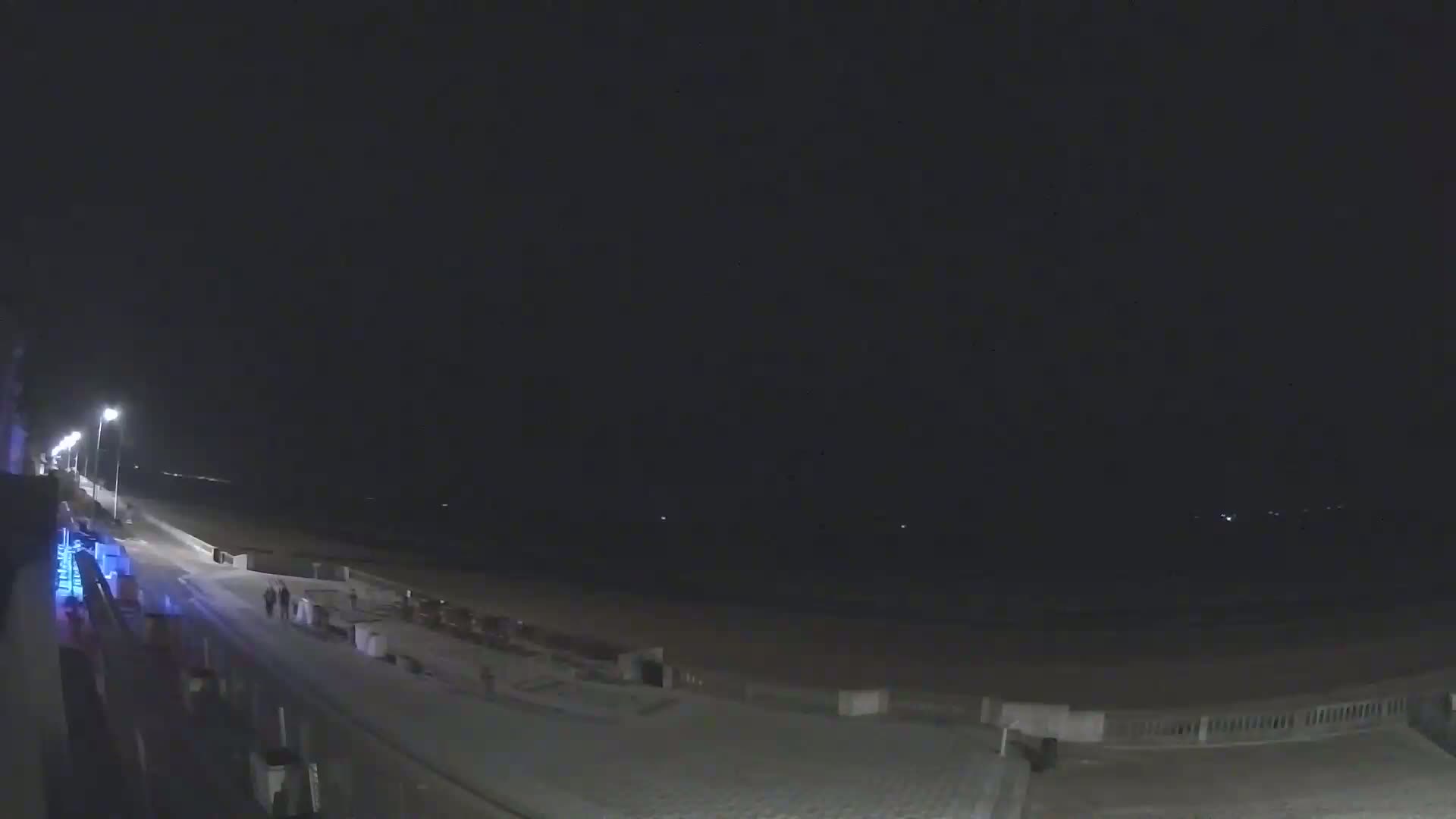 A nighttime view of a mostly empty beach boardwalk with a few people walking, lit by streetlights under a clear, dark sky.