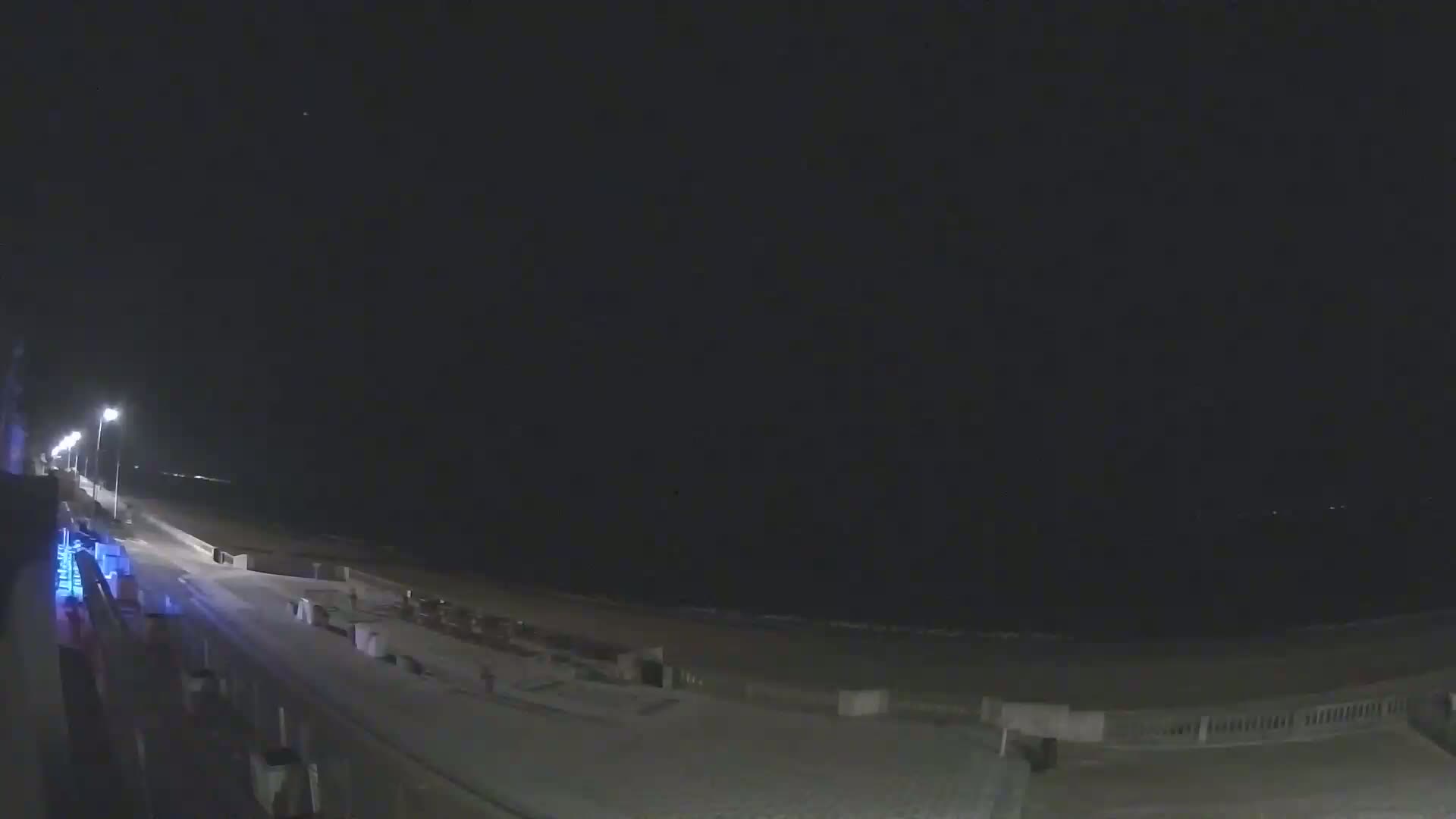 A nighttime view of a mostly empty beach with a boardwalk and streetlights, under a clear, dark sky.