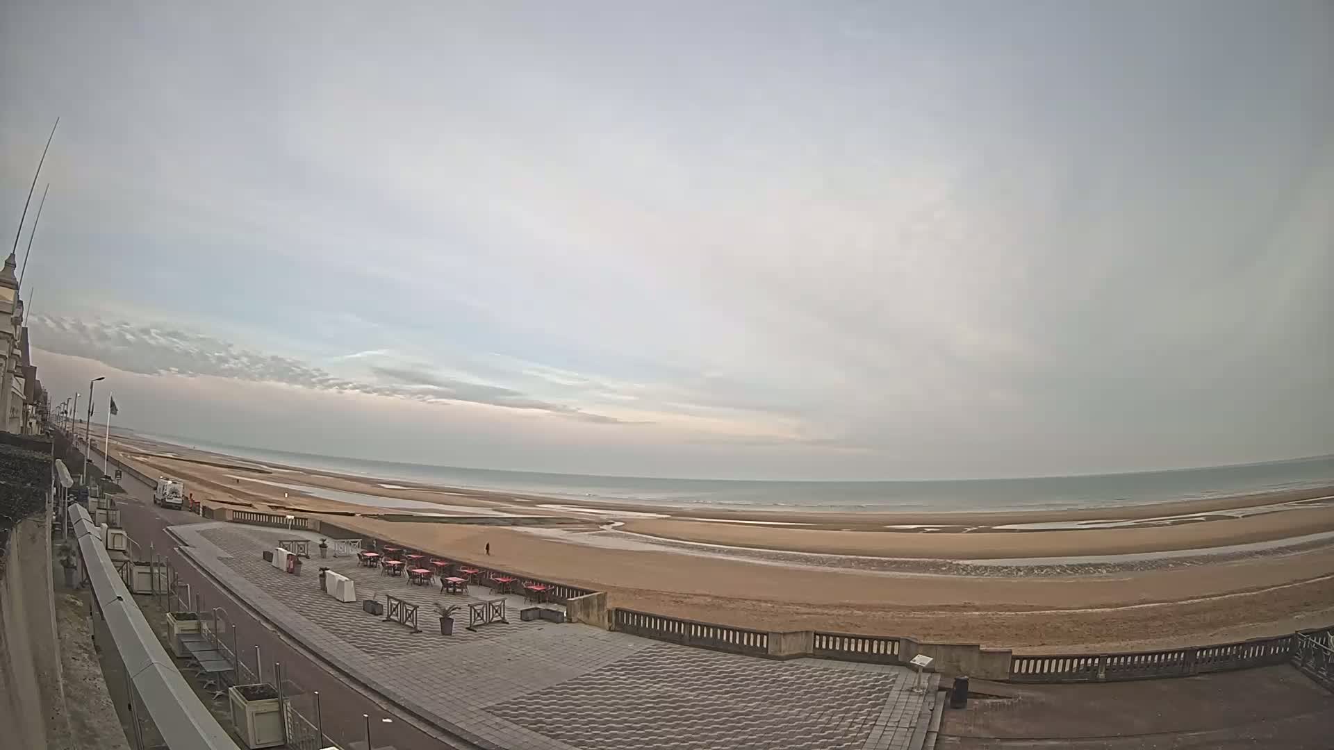 A mostly cloudy day shows an expansive beach with a calm sea,  a paved promenade with outdoor seating, and a building in the distance.