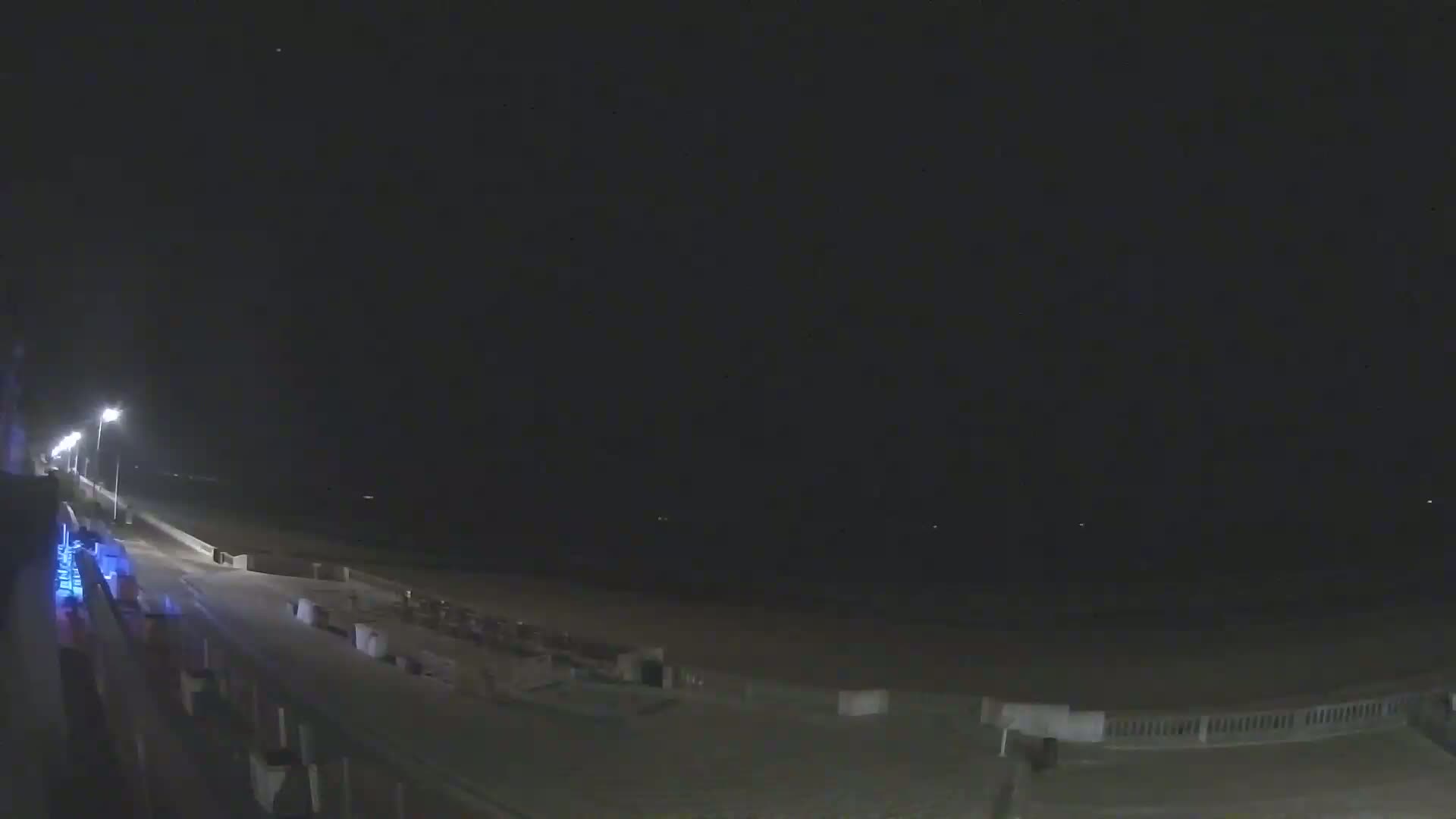 A nighttime view of a mostly empty beach with a paved walkway and dimly lit shoreline.