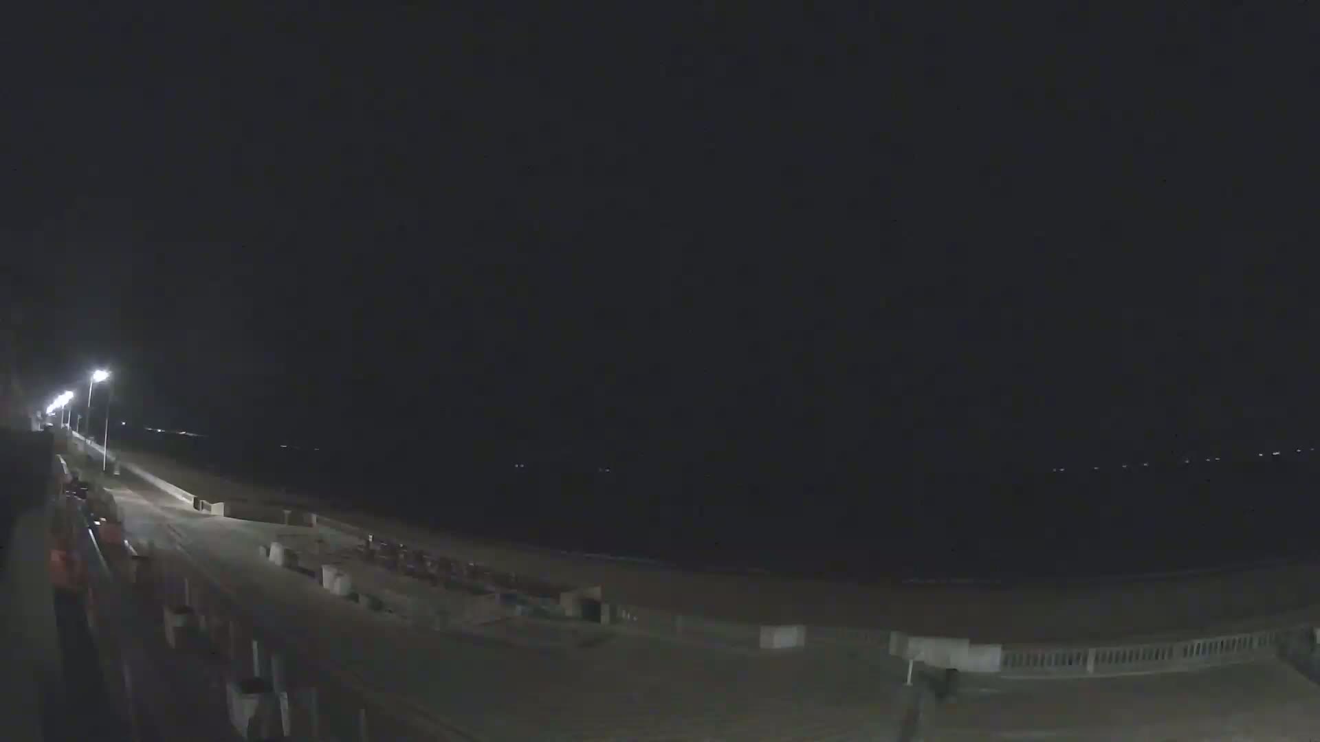 A nighttime view of a beach boardwalk illuminated by streetlights, with a dark, calm ocean in the background.