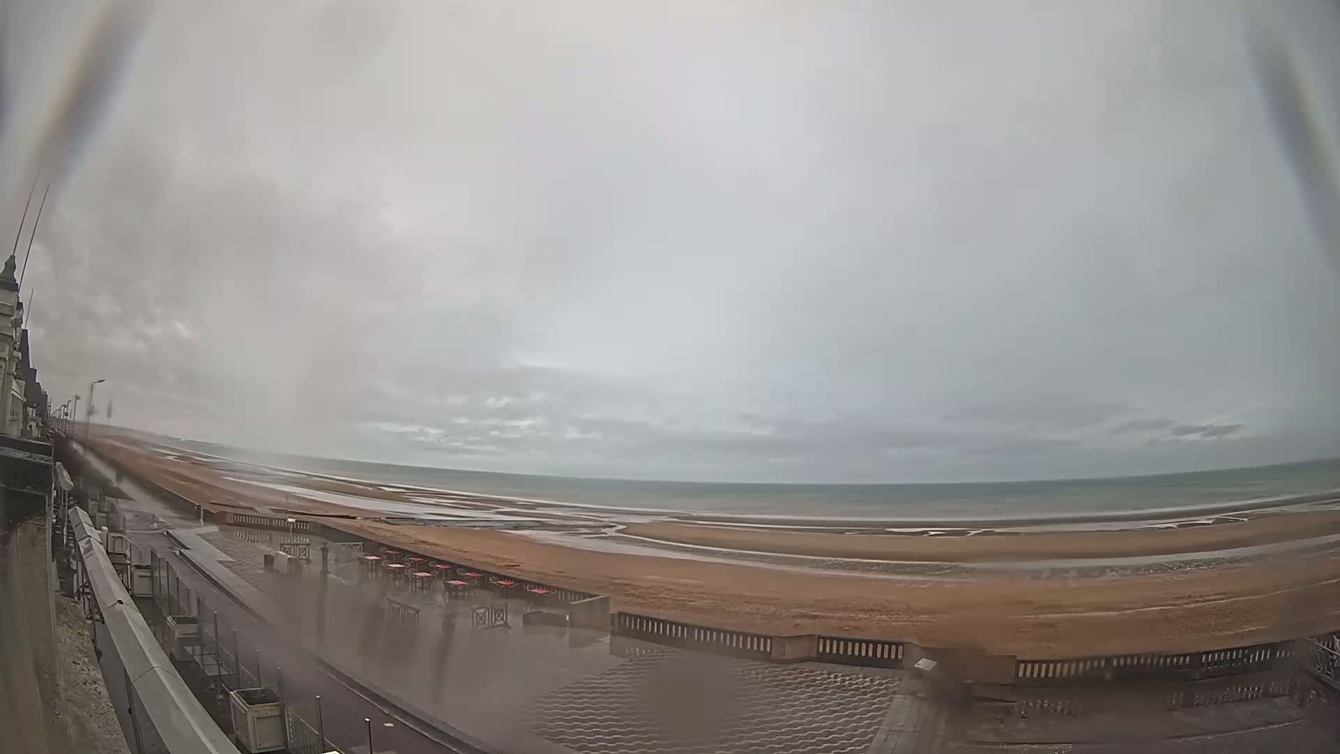 A rain-swept beach scene shows a wet sandy shore, a calm sea, and a promenade with outdoor seating, viewed from a high vantage point.