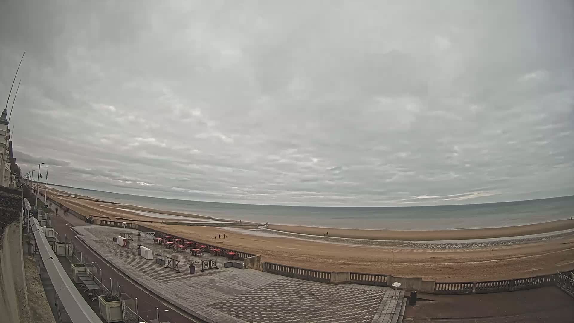 A wide shot of a beach with an overcast sky shows a mostly empty sandy beach, a paved walkway with outdoor seating, and a building in the distance.