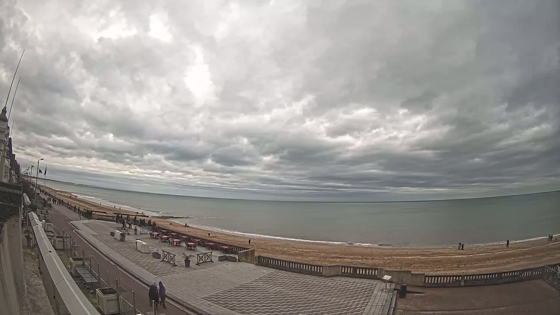 A wide, overcast sky looms over a beachside promenade with a few people strolling along a paved walkway, near outdoor seating and a sandy beach with a calm sea.