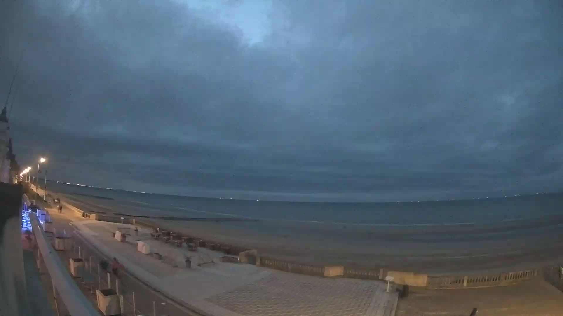 A mostly cloudy evening scene shows a beach at night with a few people walking along a paved walkway near the ocean.