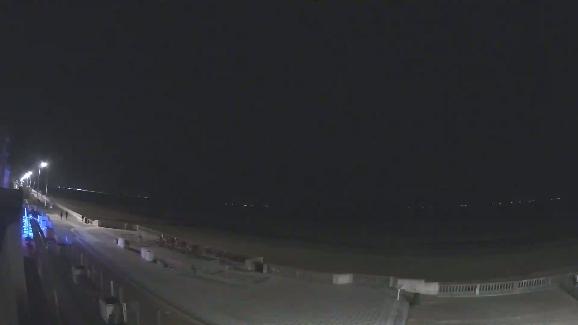 A nighttime view of a beach boardwalk with a dark sky and faint lights on the water.