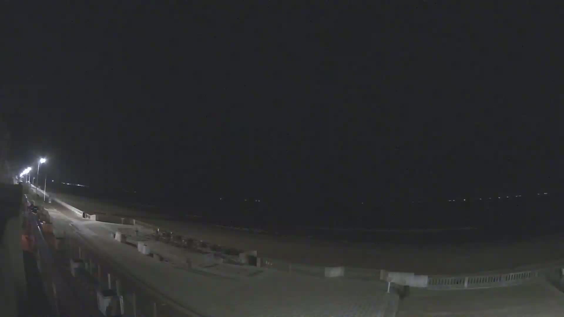 A nighttime view of a beach at night, showing a paved walkway, lights, and a dark ocean.