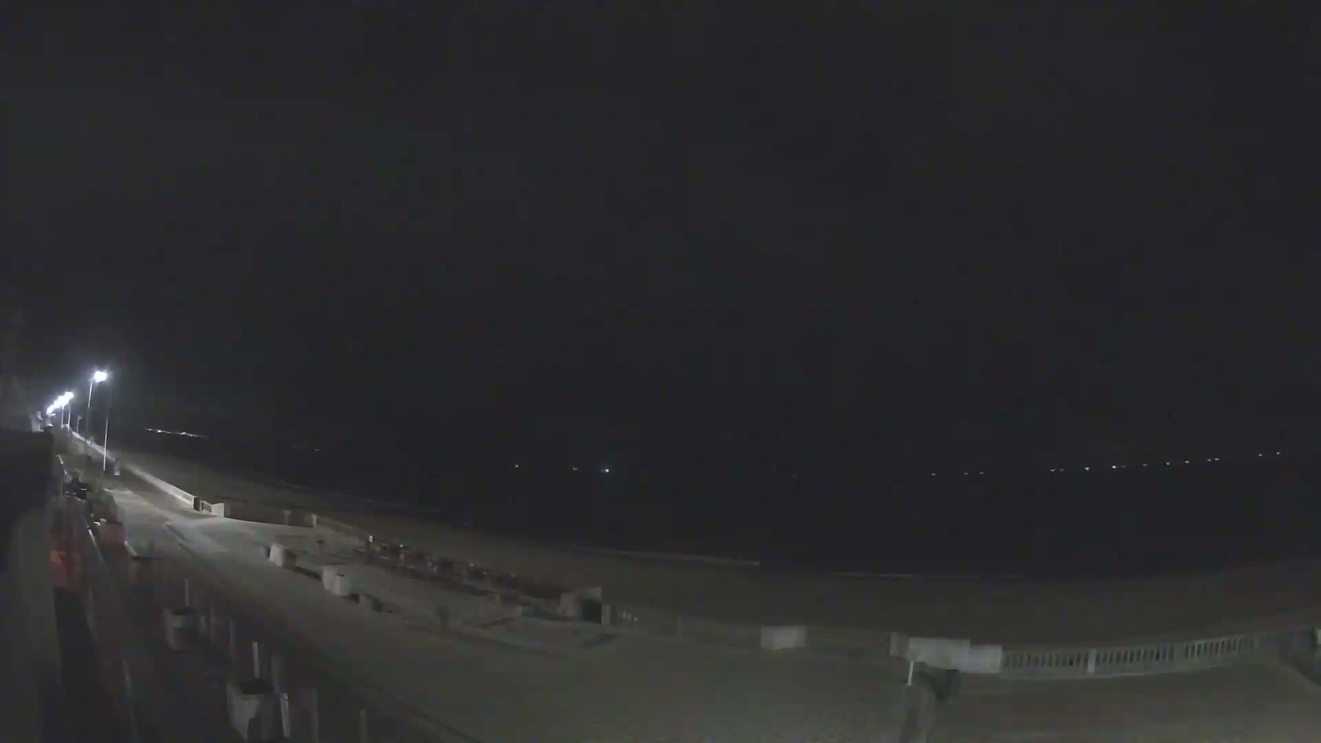 A nighttime view of a dark beach with a paved walkway lined with lights, extending toward a dark ocean with distant lights.