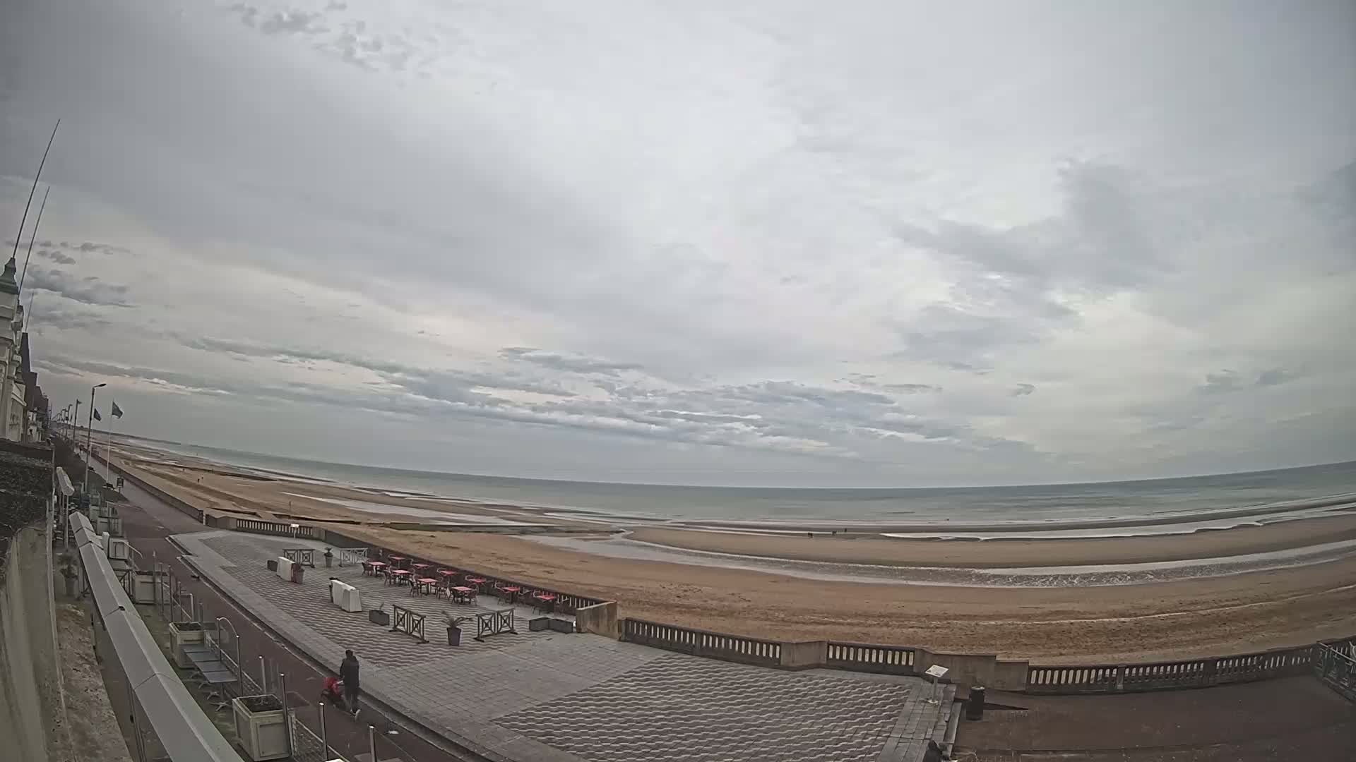 A wide shot of a beach with overcast skies, showing a paved walkway leading to a sandy beach and calm ocean waters.
