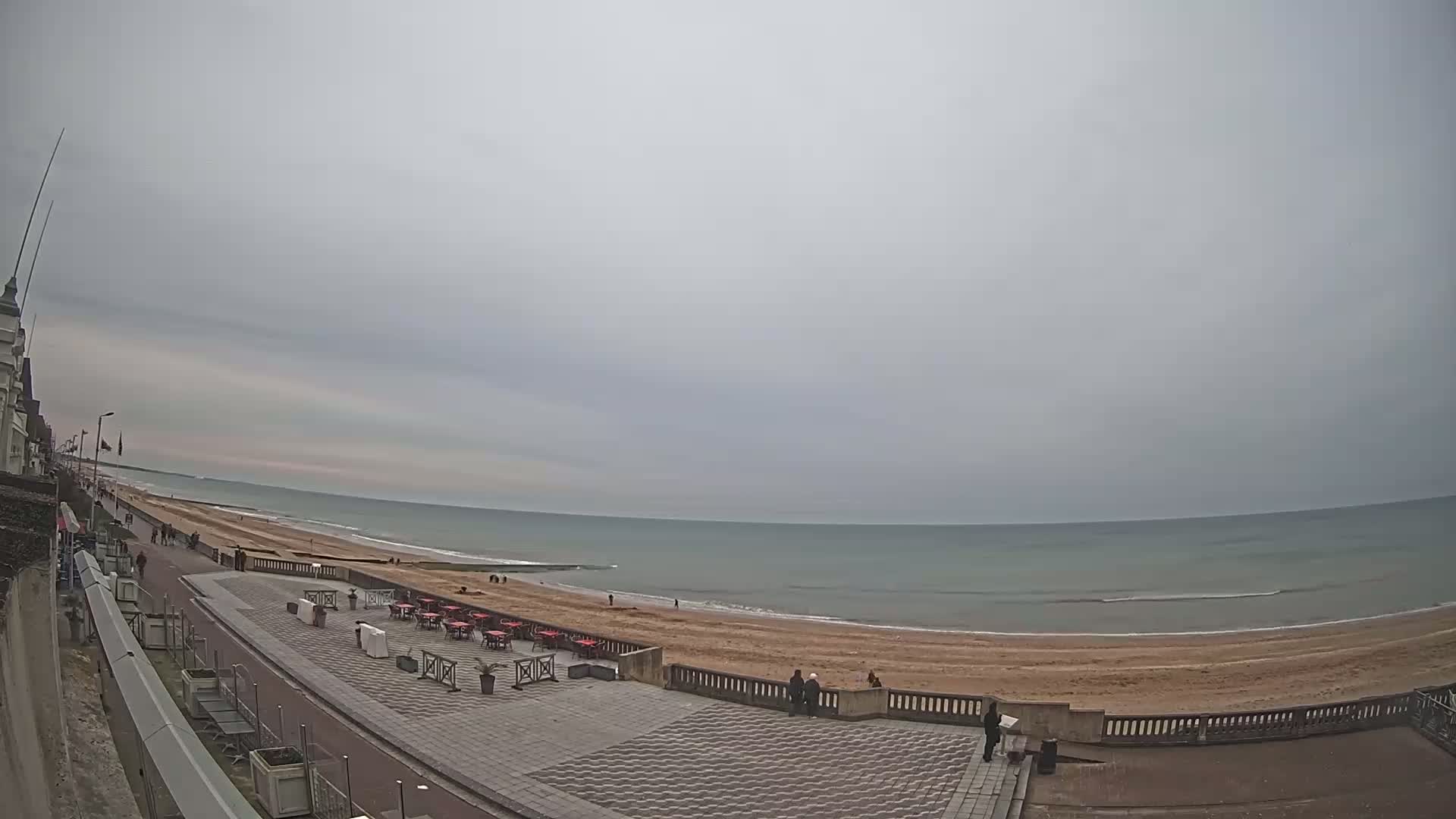 A wide, overcast beach scene shows a calm ocean, sandy beach, and a paved promenade with sparsely populated outdoor seating areas.