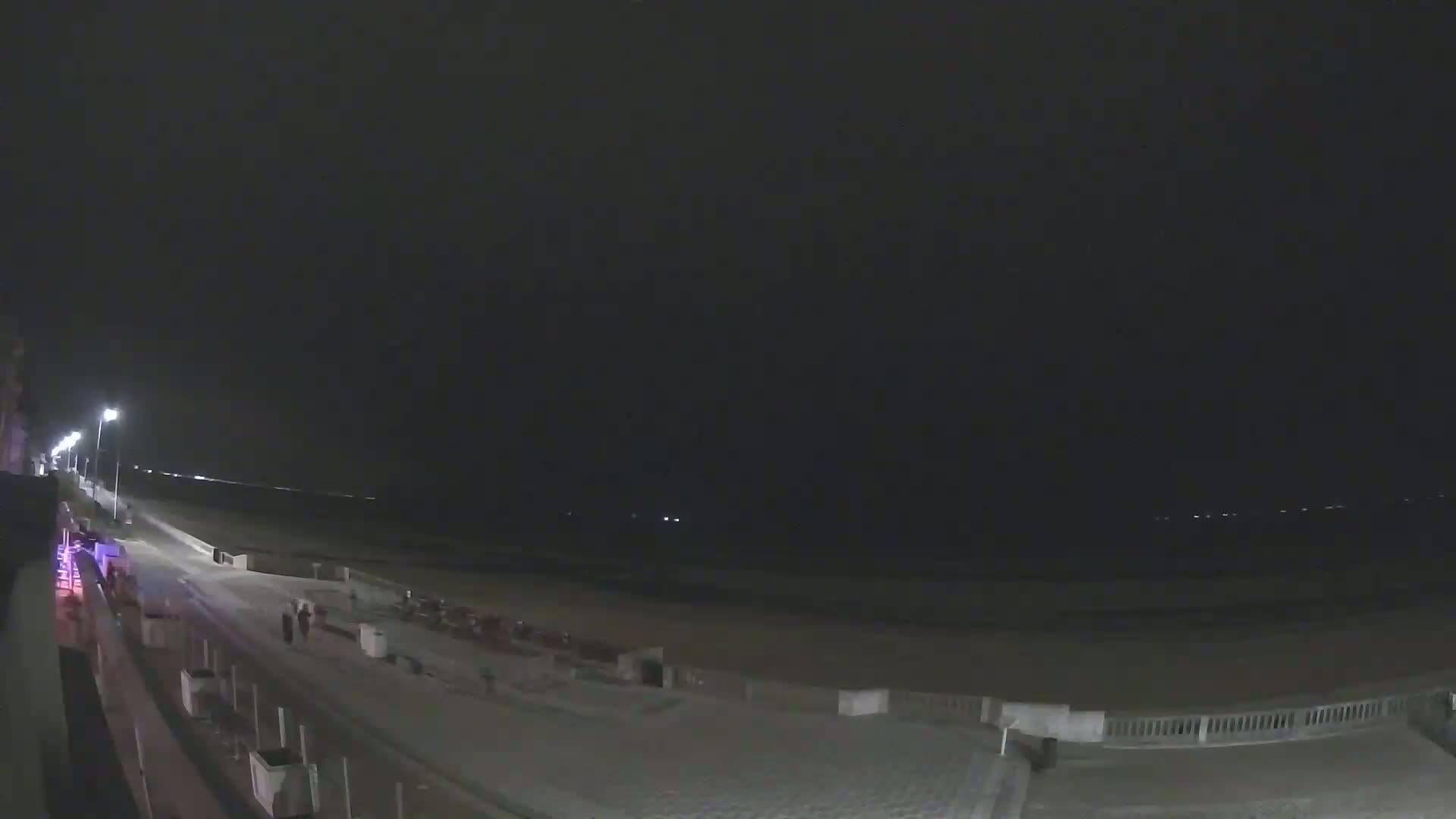 A nighttime view of a mostly empty beach with a boardwalk and a few people visible under a dark sky.