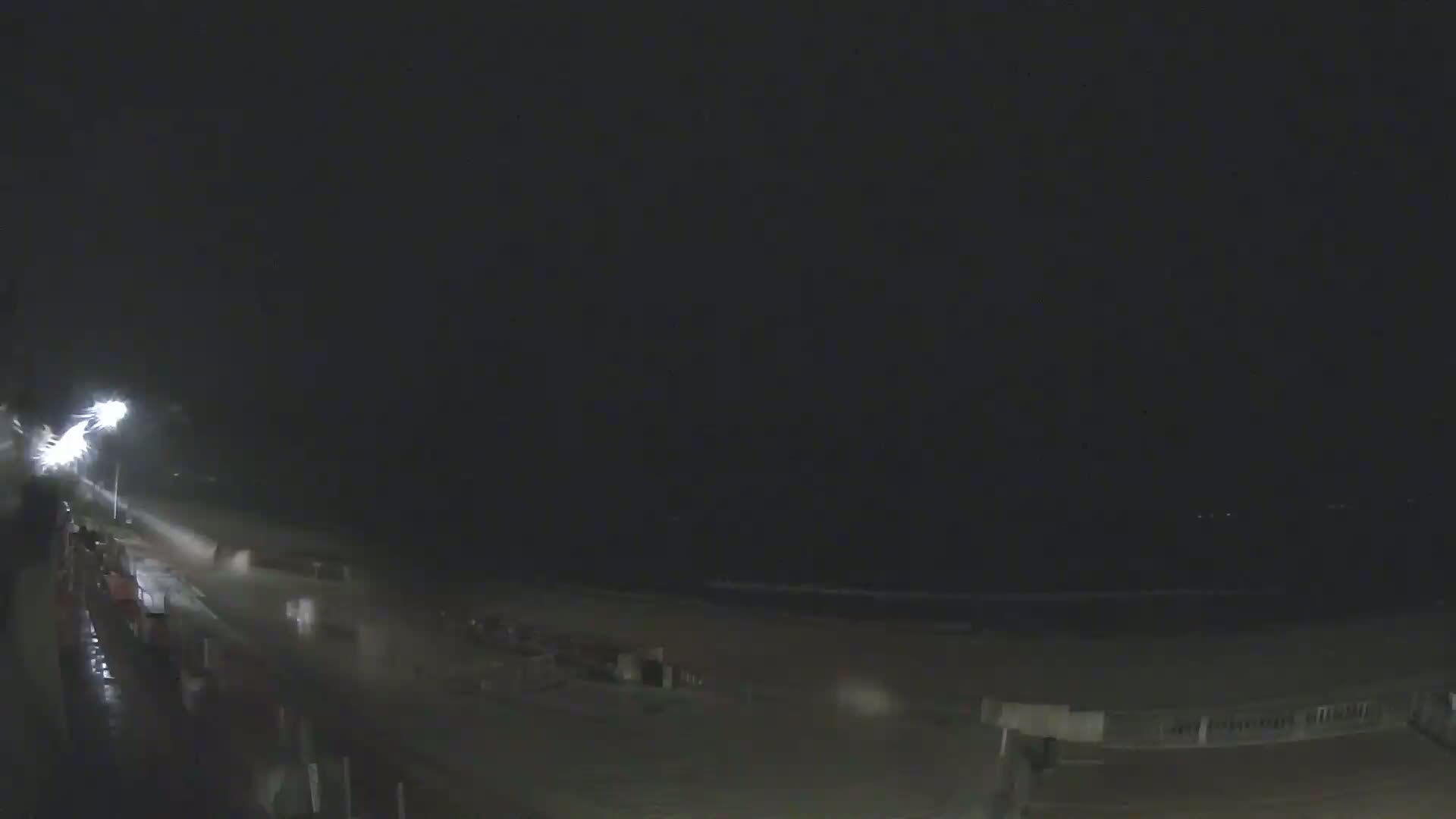 A nighttime view of a rain-swept beach, with a brightly lit walkway and ocean visible in the distance.