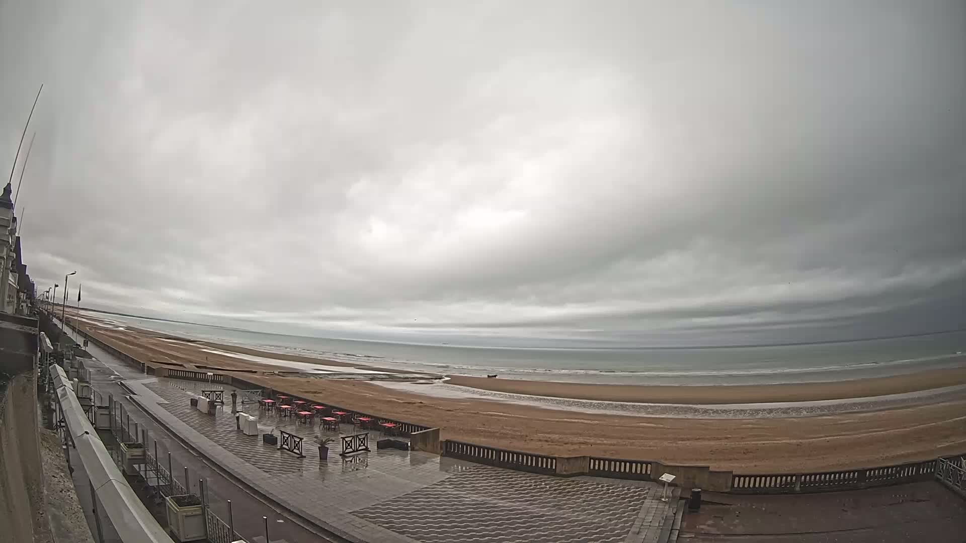 A wet, paved seaside promenade overlooks a wide sandy beach and calm ocean under an overcast sky.