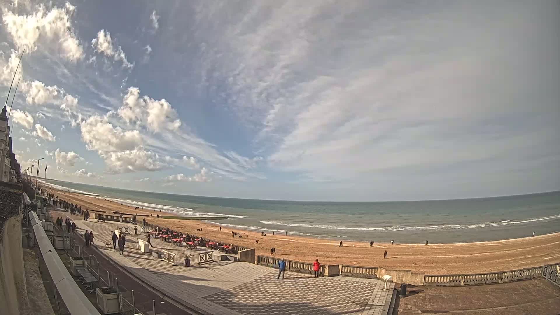 A partly cloudy day at a beach shows a wide expanse of ocean, a sandy shore with people walking, and a paved walkway with outdoor seating near a building.