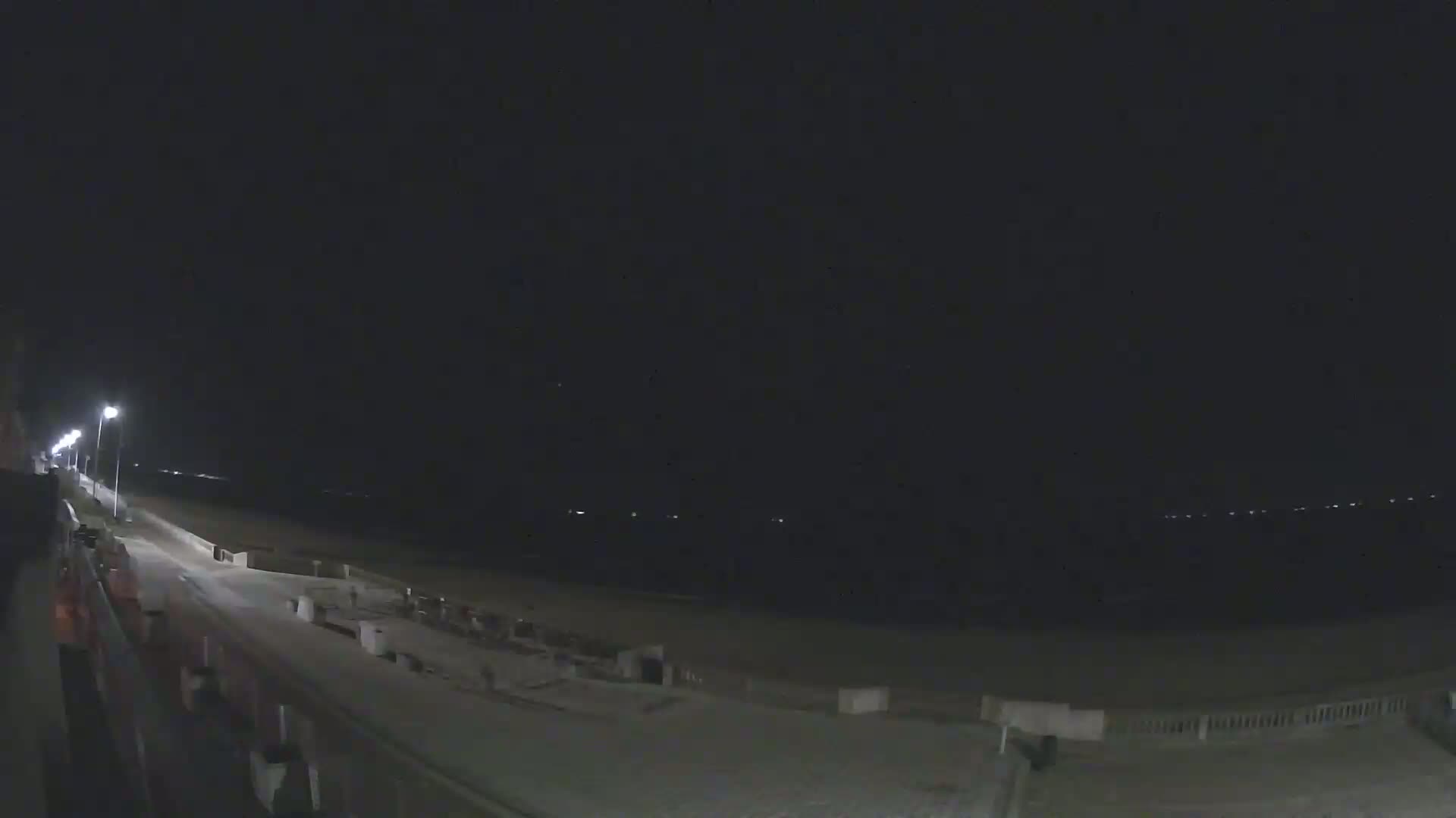 A nighttime view of a beach at night, showing a paved walkway, lights, and a dark ocean.
