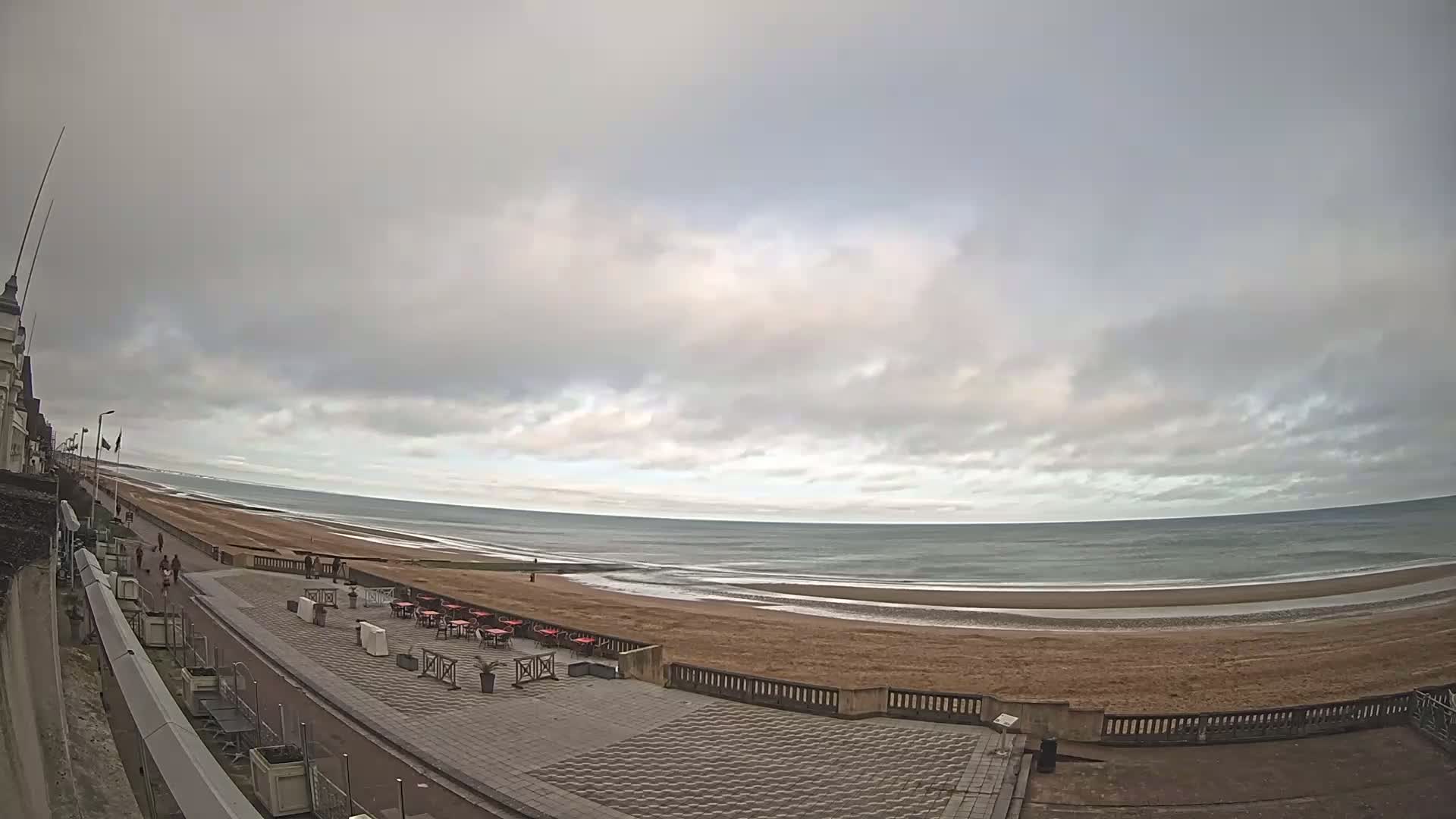 A paved beachfront promenade with outdoor seating overlooks a wide, sandy beach and a calm, overcast ocean.