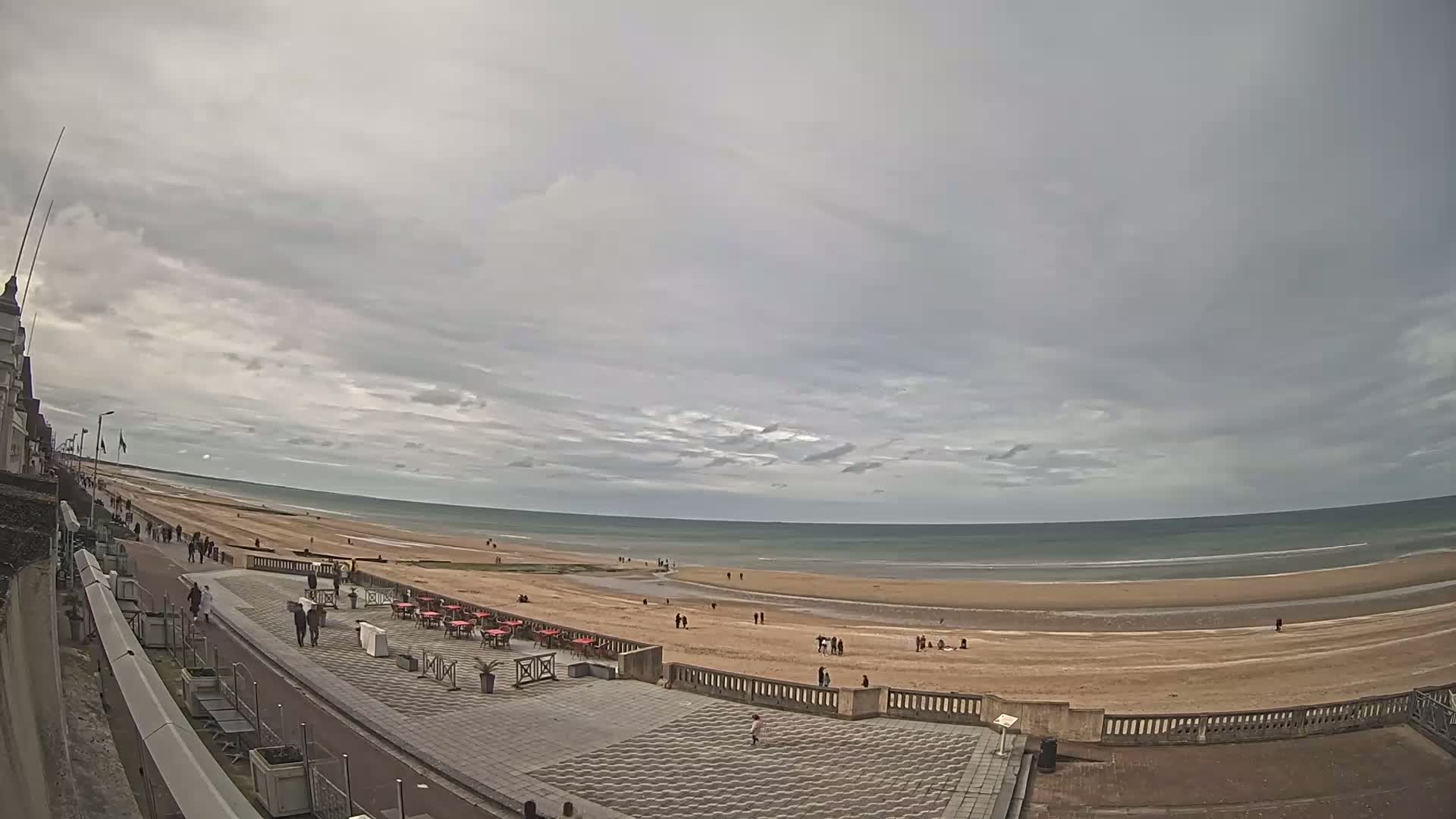 A wide, overcast sky overlooks a long, sandy beach with a few people scattered along it and a promenade with outdoor seating.