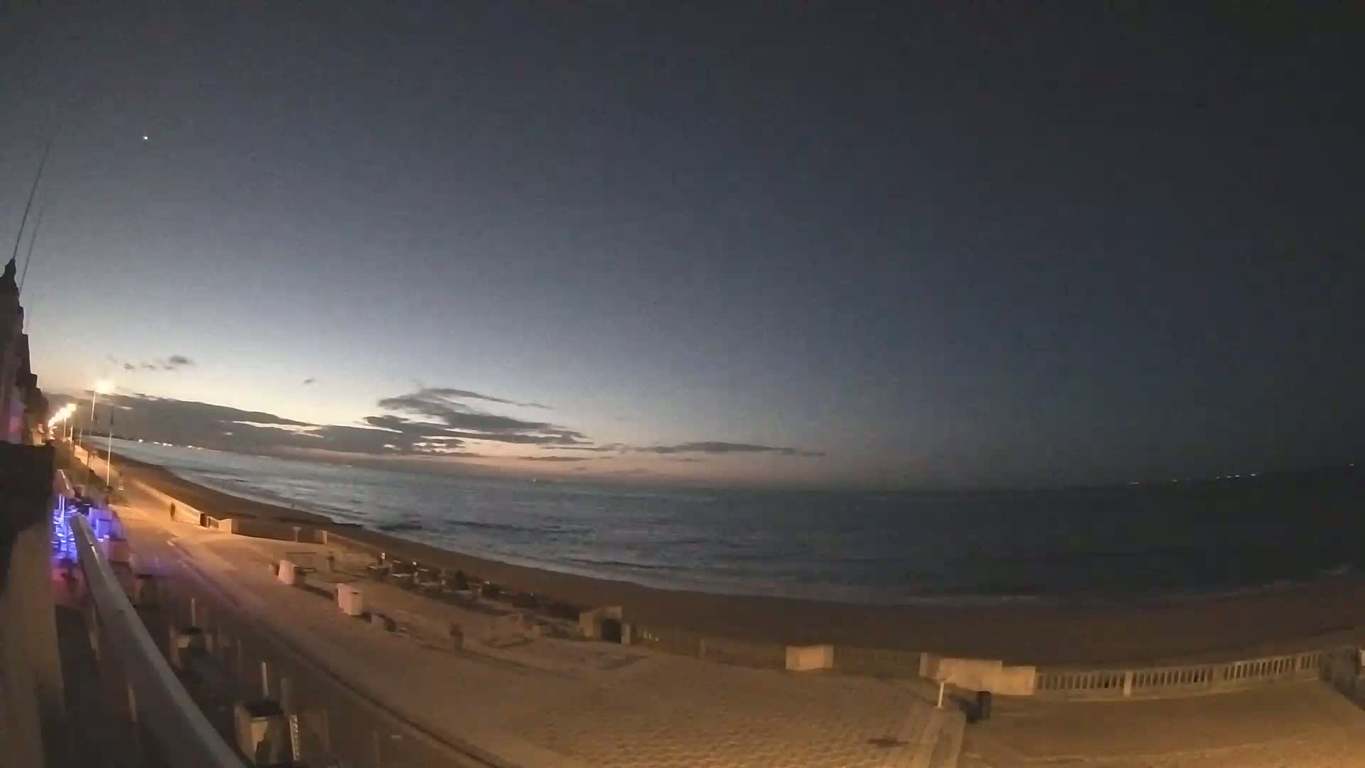 A dark, pre-dawn beach scene shows a calm ocean under a mostly clear, twilight sky.