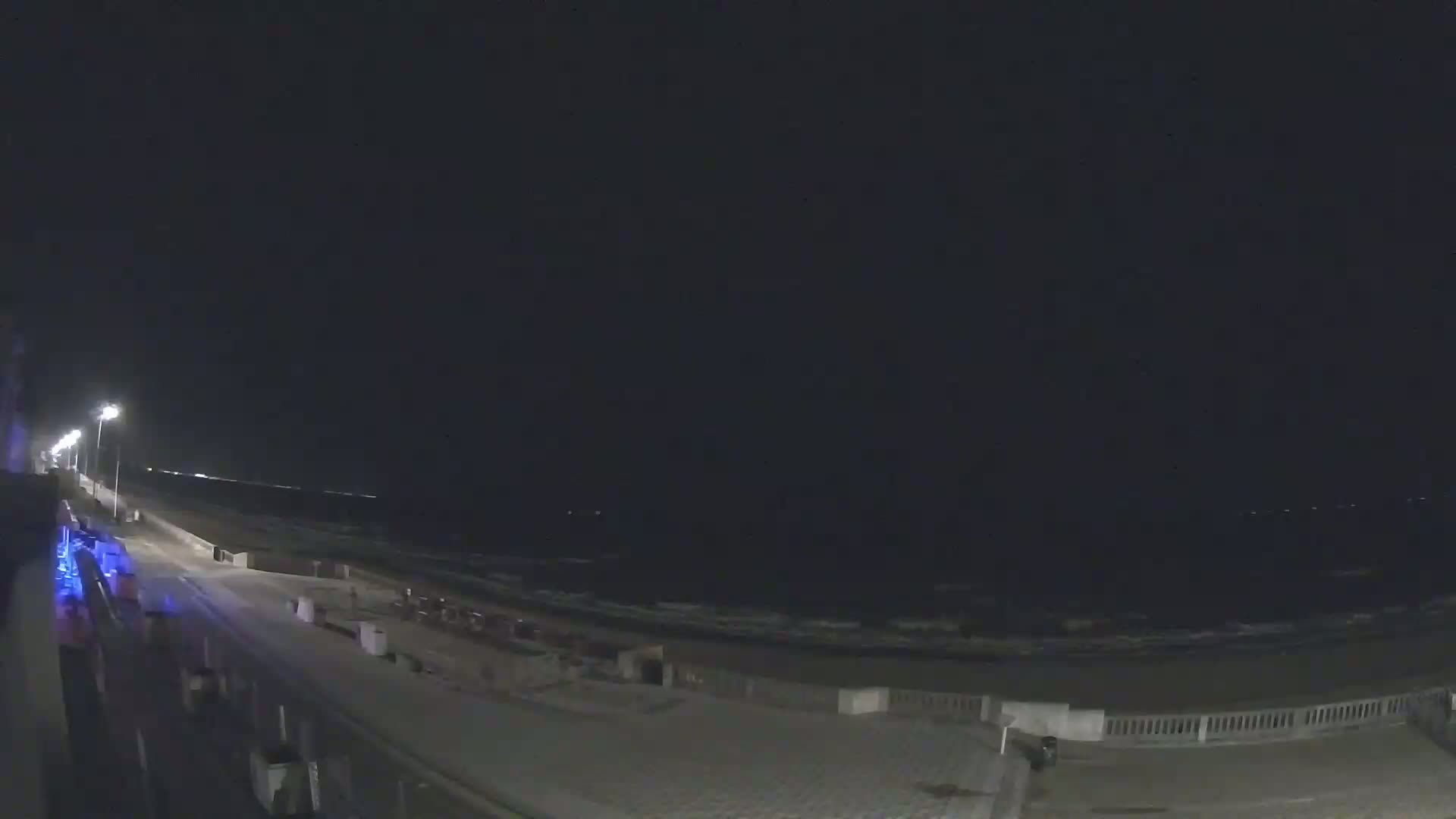 A nighttime view of a beach with gentle waves under a dark sky, showing a paved walkway and low lighting along the shoreline.