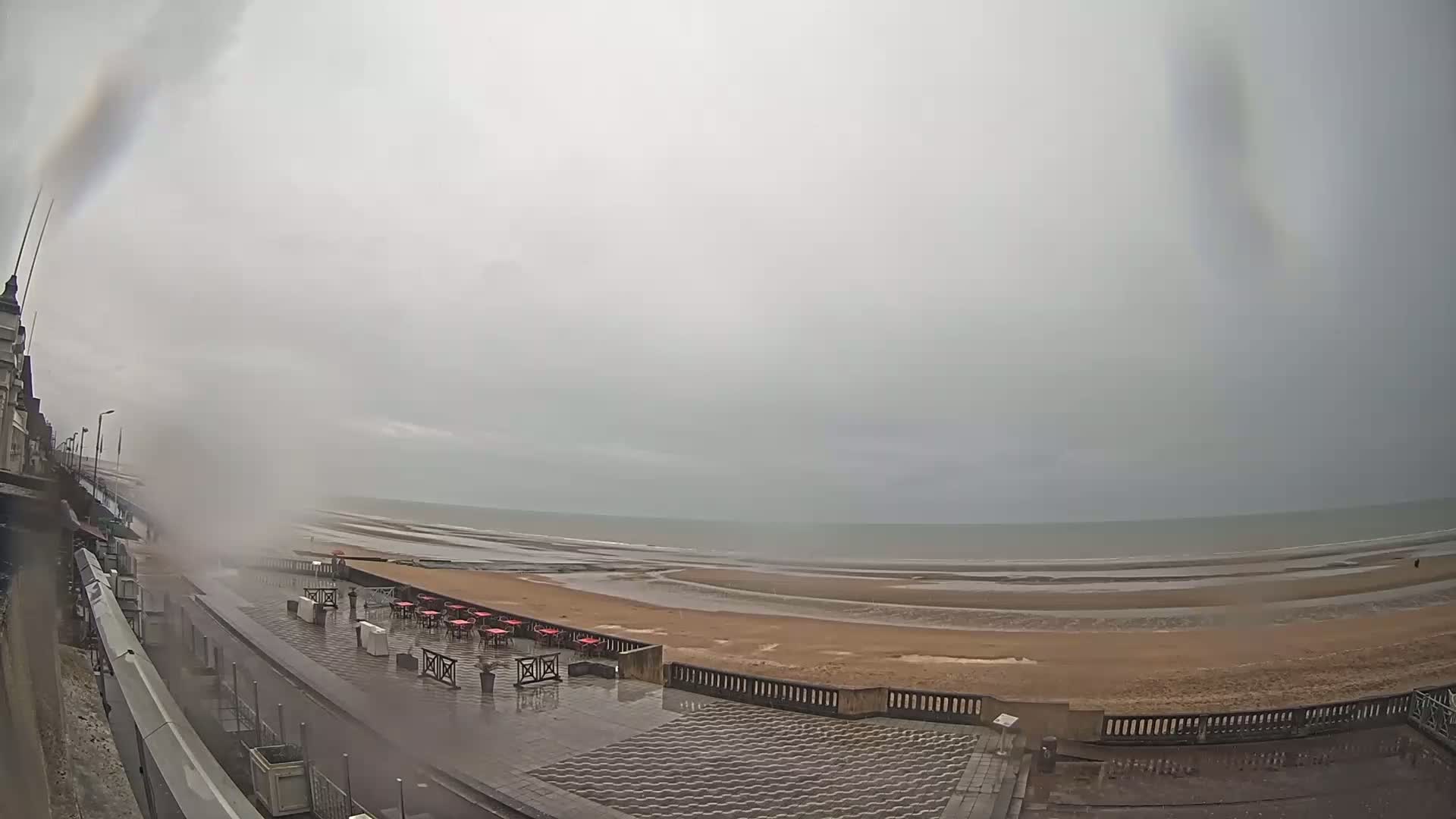 A wet, overcast day at a seaside promenade shows a beach with a low tide, a row of outdoor tables and chairs, and a building in the background.