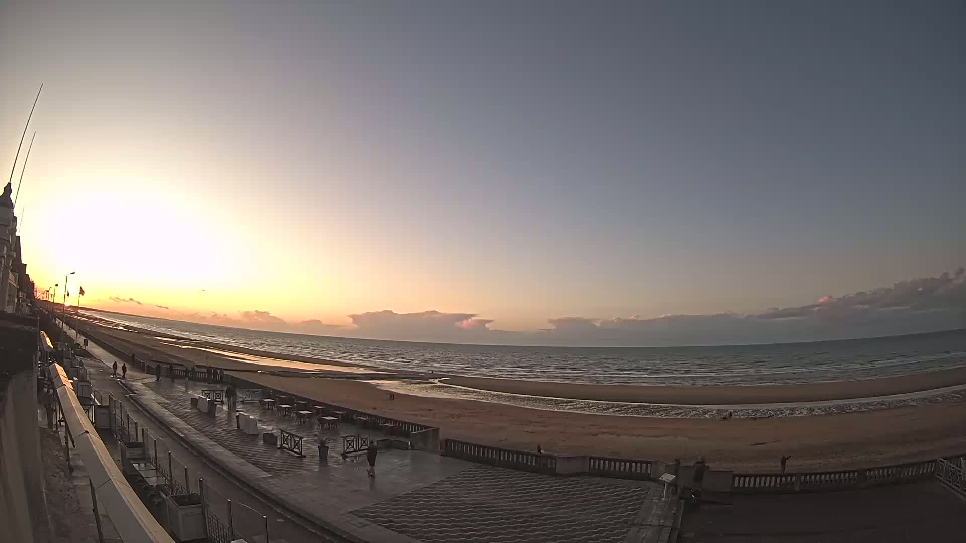 A wide shot shows a beach at sunset, with a paved walkway and a few people visible, under a mostly clear sky with some clouds on the horizon.
