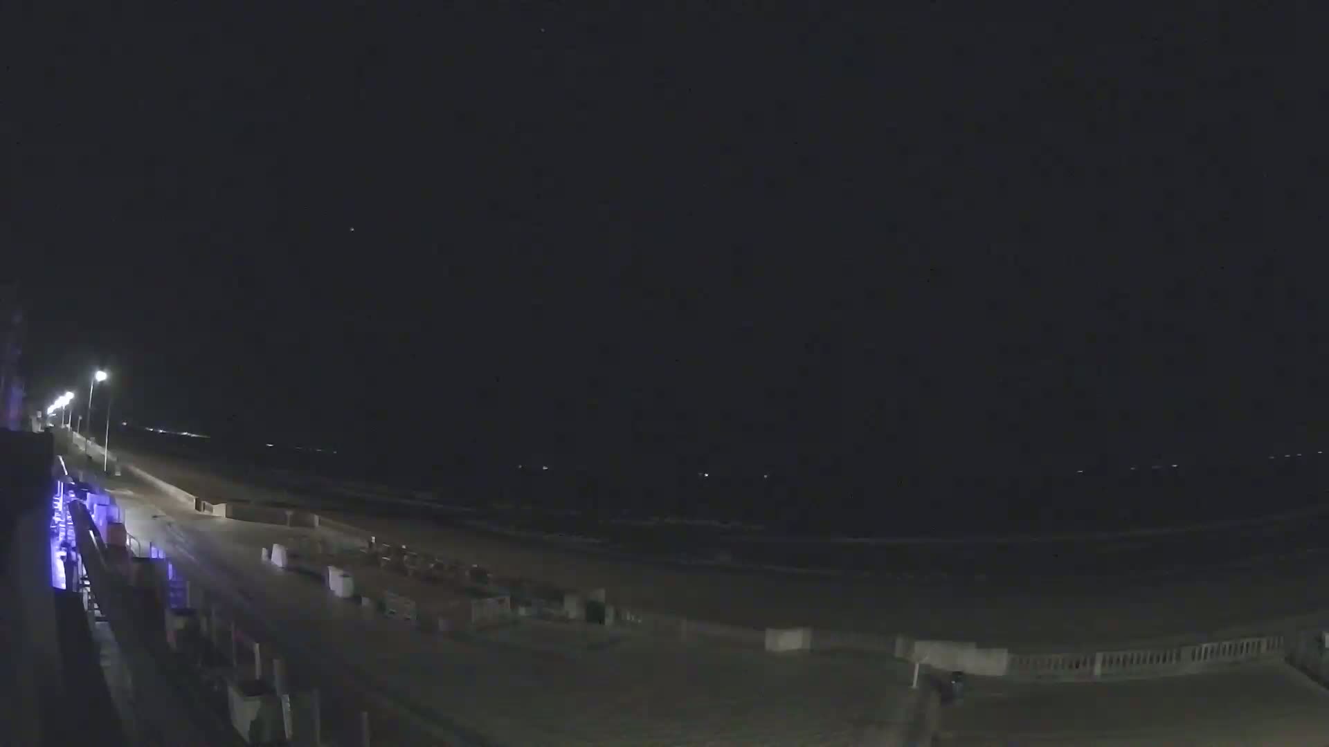 A nighttime view of a mostly empty beach boardwalk with dimly lit ocean waves in the background.