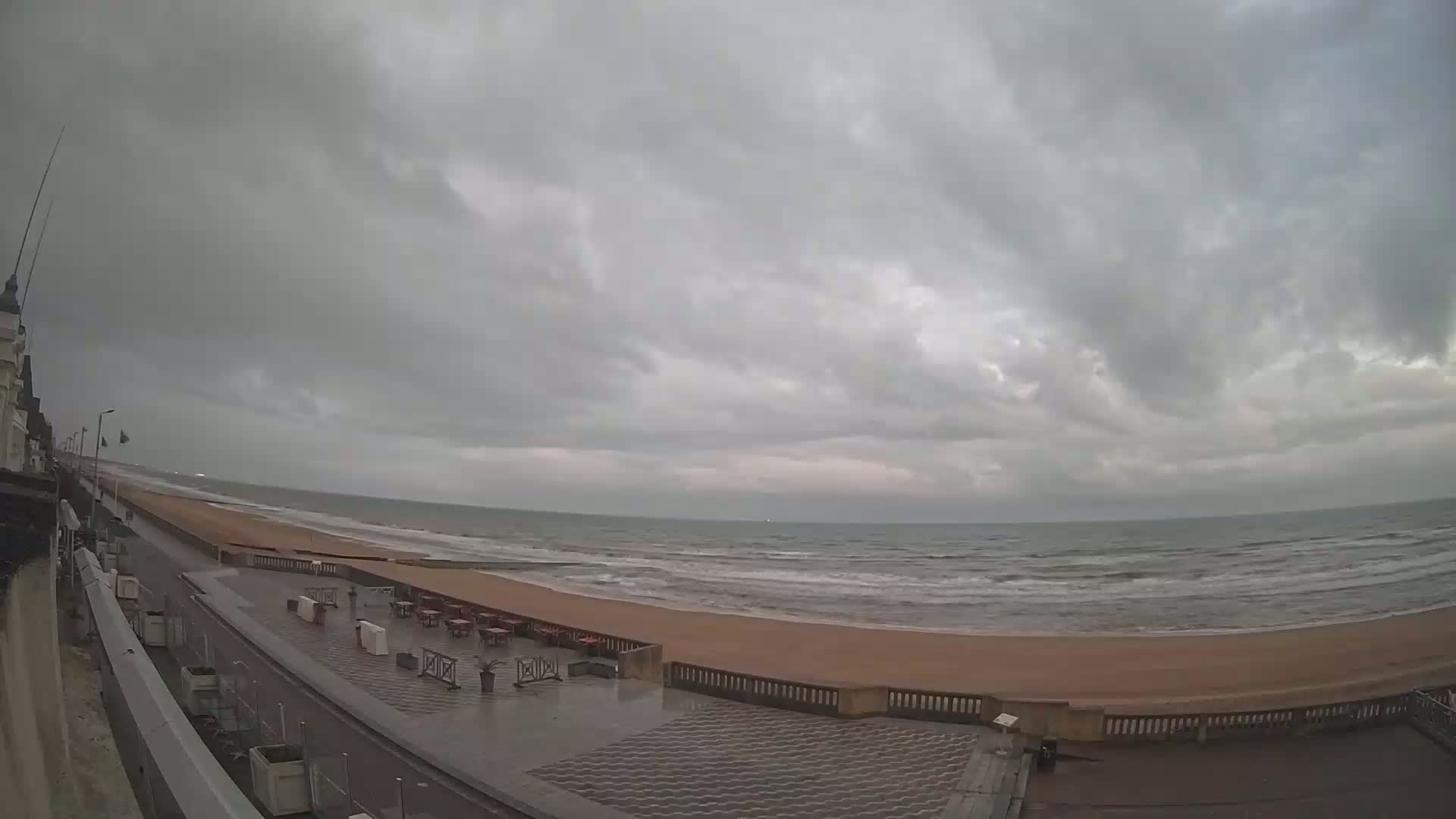 A mostly cloudy day shows a wide sandy beach with a calm sea and a paved walkway lined with outdoor seating, all under a gray sky.