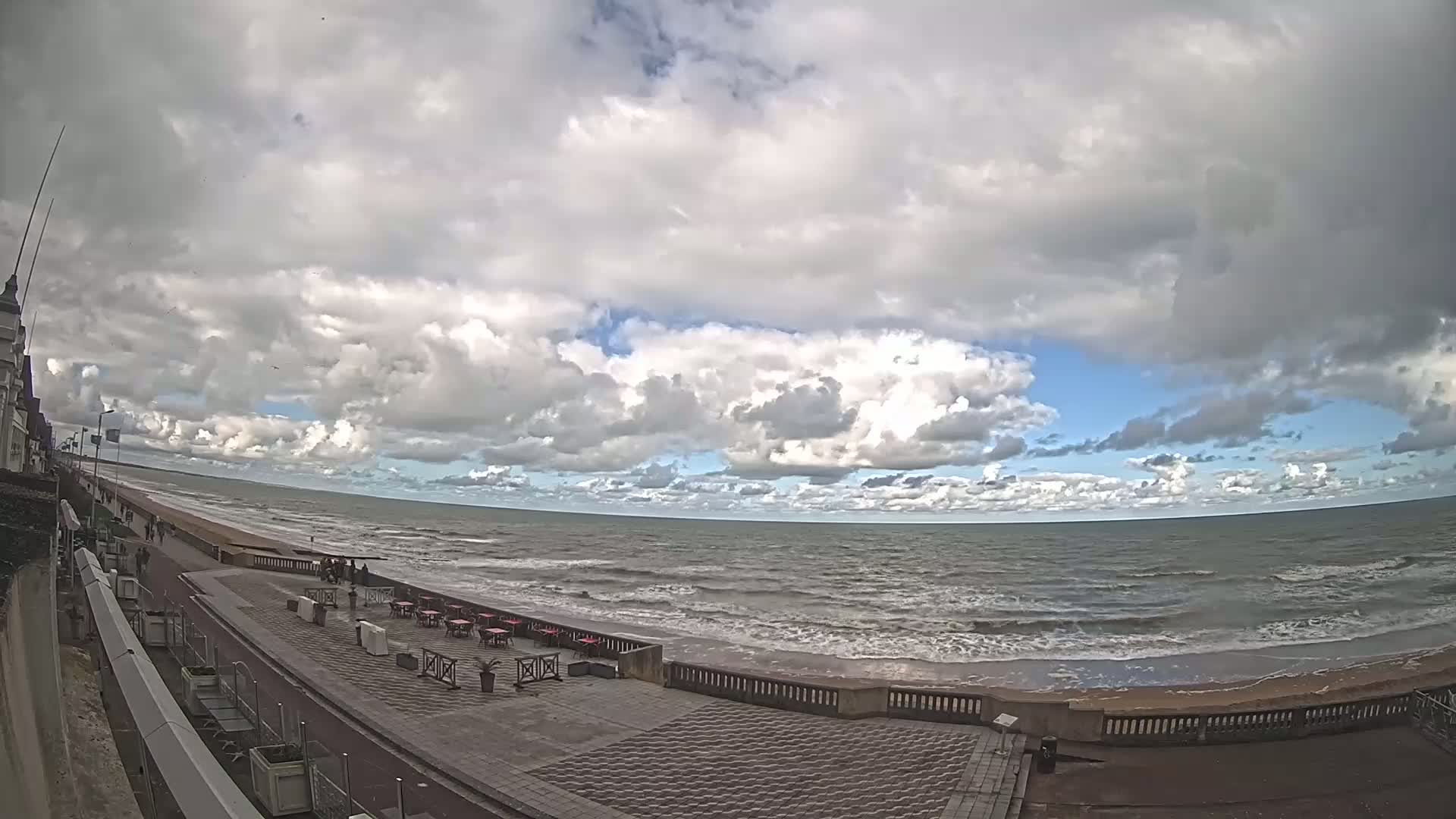 A partly cloudy day shows a mostly calm ocean with small waves lapping a sandy beach, along which a paved promenade with outdoor seating stretches beside a building.