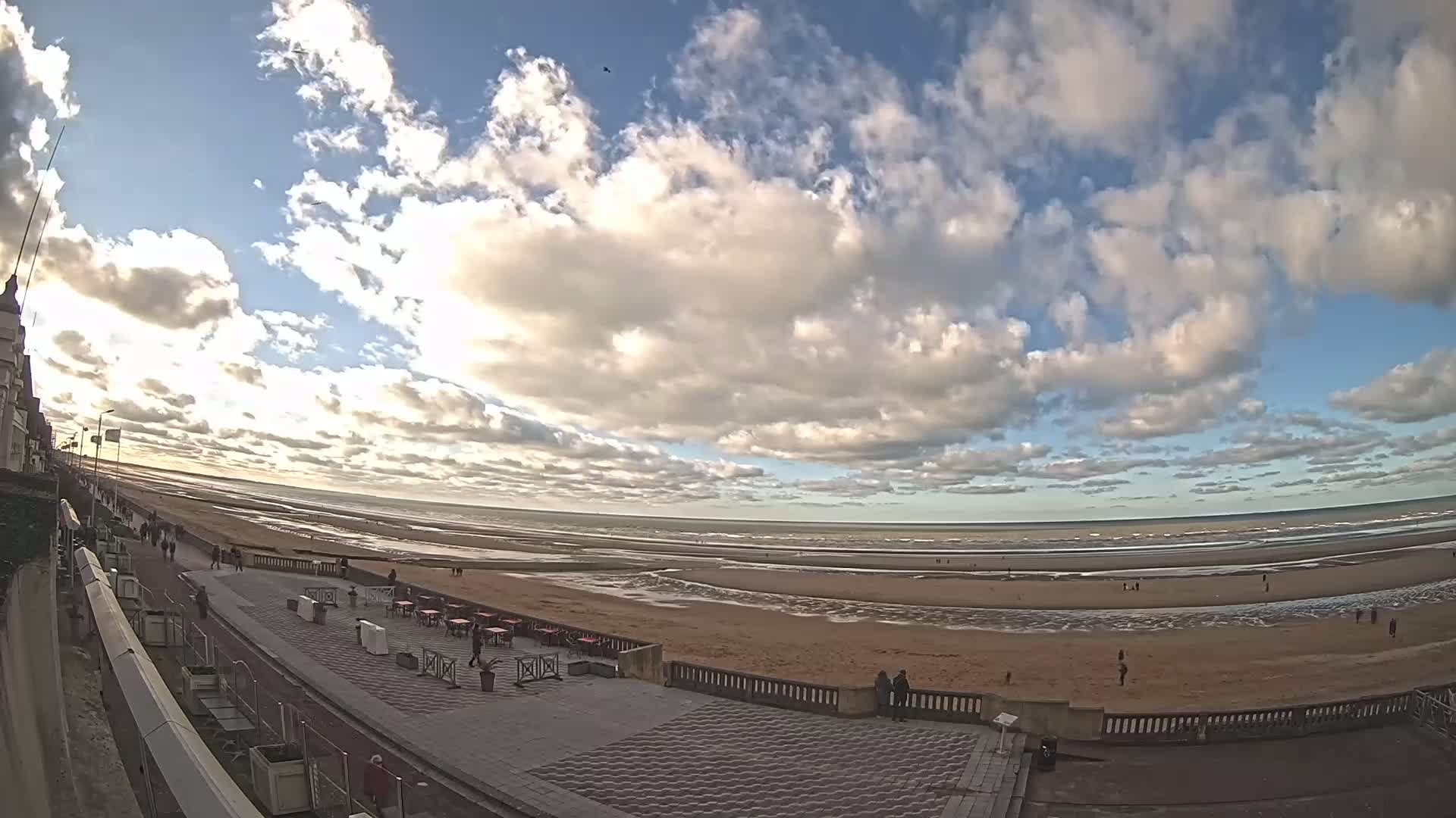 A wide, partly cloudy sky overlooks a beach with a paved walkway and several outdoor tables, with people scattered along the shore and promenade.