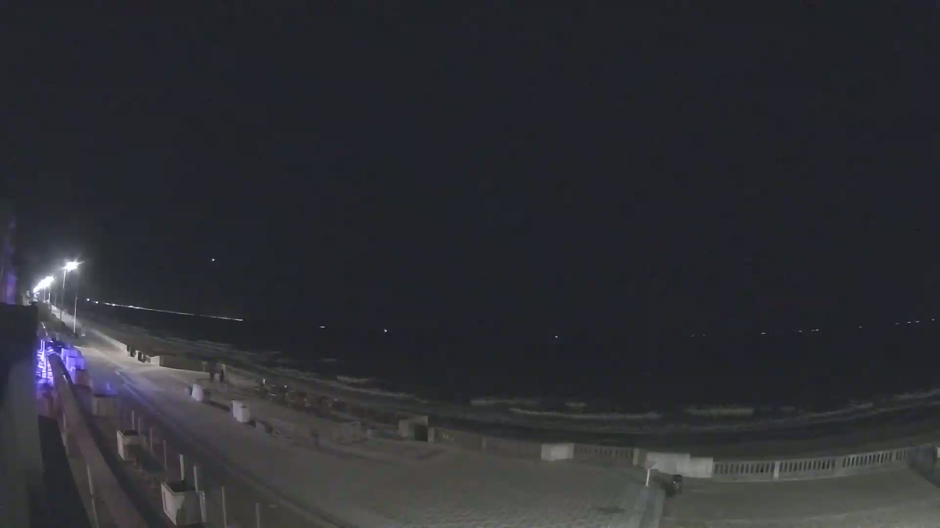 A nighttime view of a beach with gentle waves under a dark, clear sky, showing a paved walkway and some streetlights.