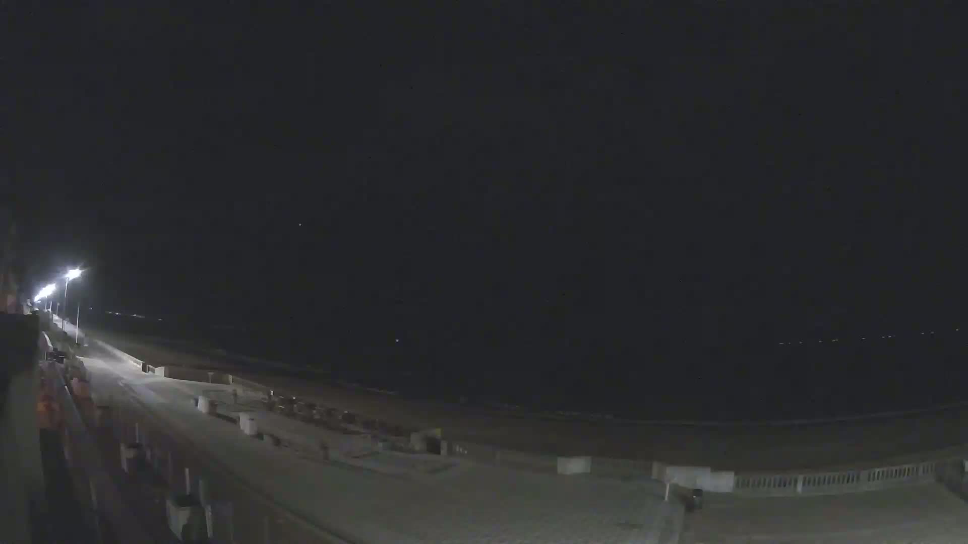 A nighttime view of a beach boardwalk illuminated by streetlights, with a dark ocean in the background under a clear sky.