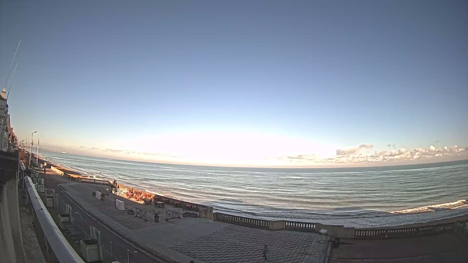A mostly clear sky overlooks a calm ocean meeting a paved beachfront promenade with a few people and outdoor seating.