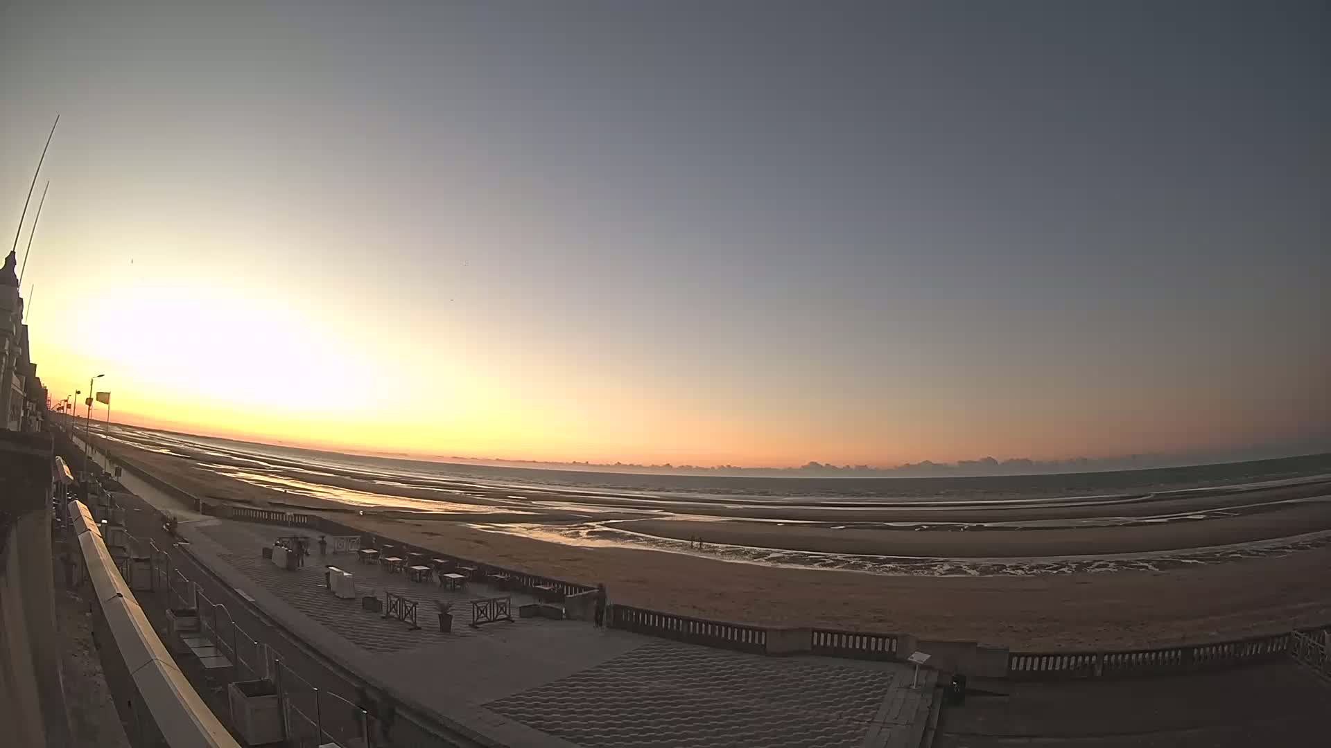 A wide shot of a beach at sunset, showing a boardwalk, ocean, and some outdoor seating areas.